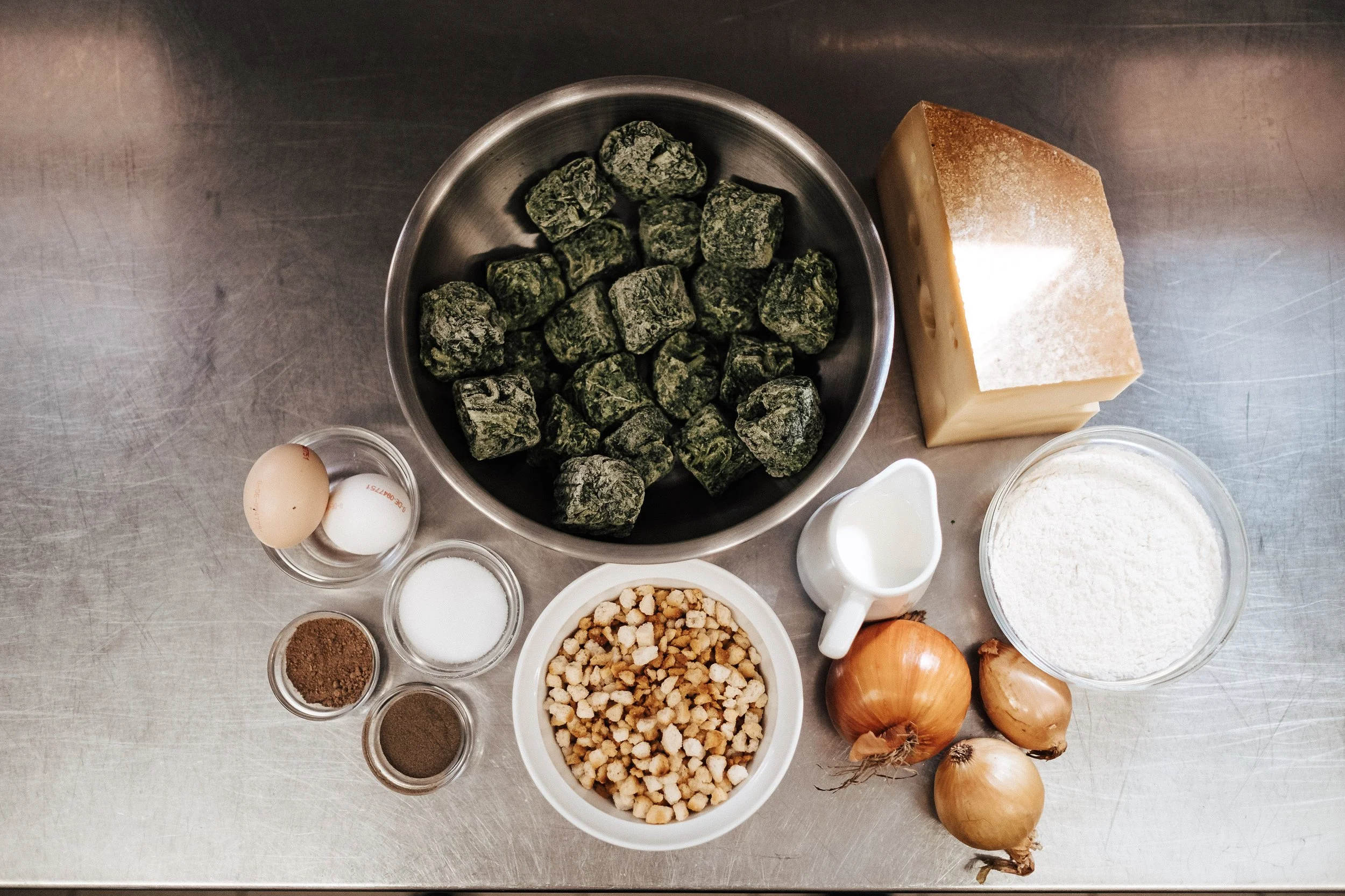 Verschiedene Zutaten für ein Rezept, darunter grünes Meersalz, Parmesan-Käse, Eier, Zwiebeln, Knoblauch, Mehl, und Gewürze, auf einem metallischen Tisch.