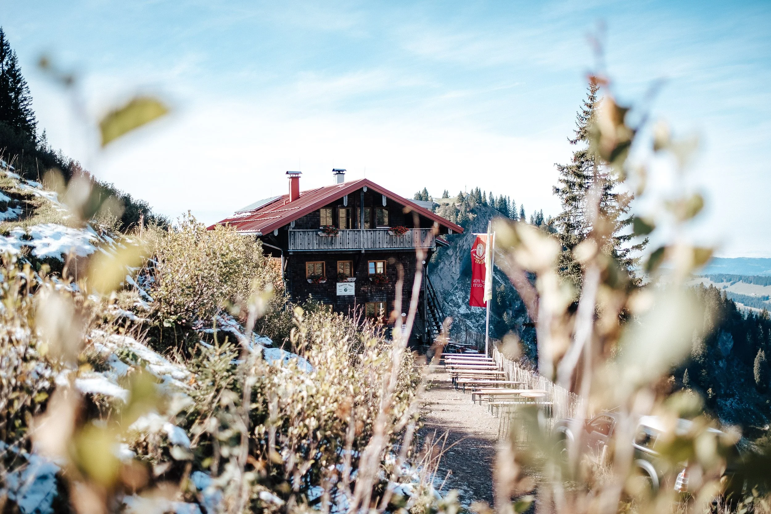 Ein Chalet in den Bergen mit roten Dach und Außenterrasse, umgeben von Schneeresten und kahlen Büschen, im Hintergrund Tannen und Hügel bei blauem Himmel.