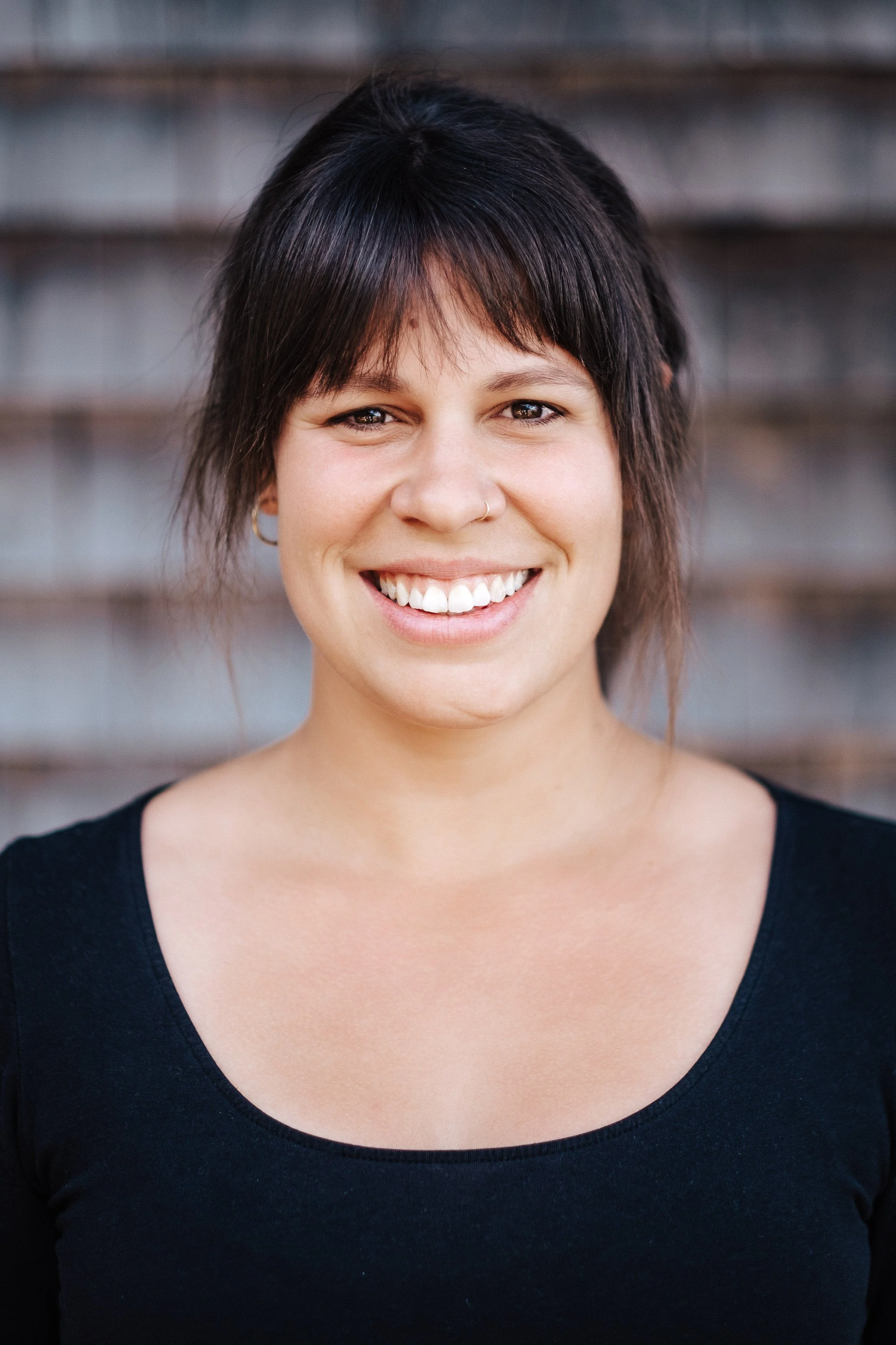 Porträt einer lachenden Frau mit dunklem Haar, Stern-Nase und schwarzem Oberteil vor einer grauen Steinmauer.