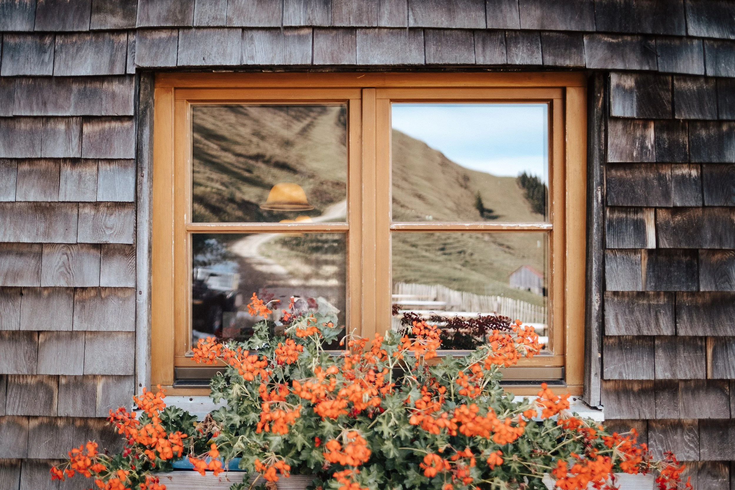 Holzverkleidetes Fenster mit Medaillon und orange Blumen vor einer ländlichen Berglandschaft.