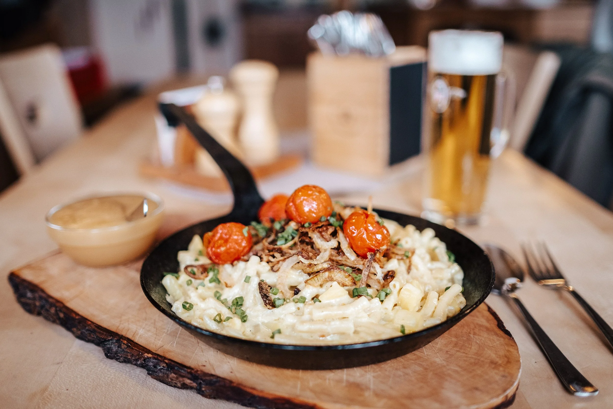 Käsespätzle in einer Pfanne mit gerösteten Zwiebeln und roten Oliven, dazu ein Glas Bier und eine Soße, serviert auf einem Holzbrett auf einem Tisch in einem Restaurant.