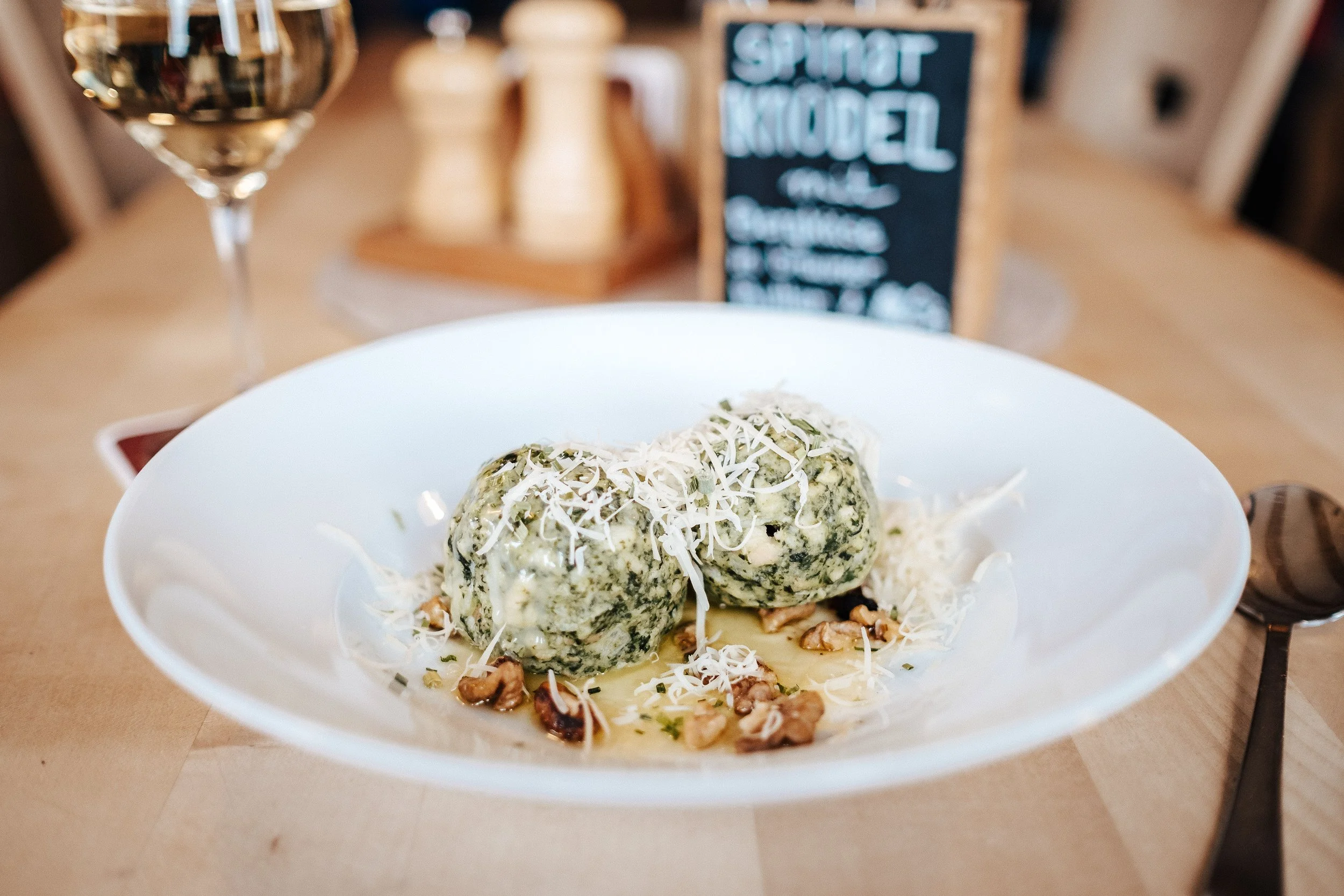 Frikadellen mit Käse, serviert auf einem weißen Teller, mit geriebenem Käse, Walnüssen und einer cremigen Soße, im Hintergrund ein Glas Weißwein und ein Schild mit der Aufschrift 'Spinatknödel'.