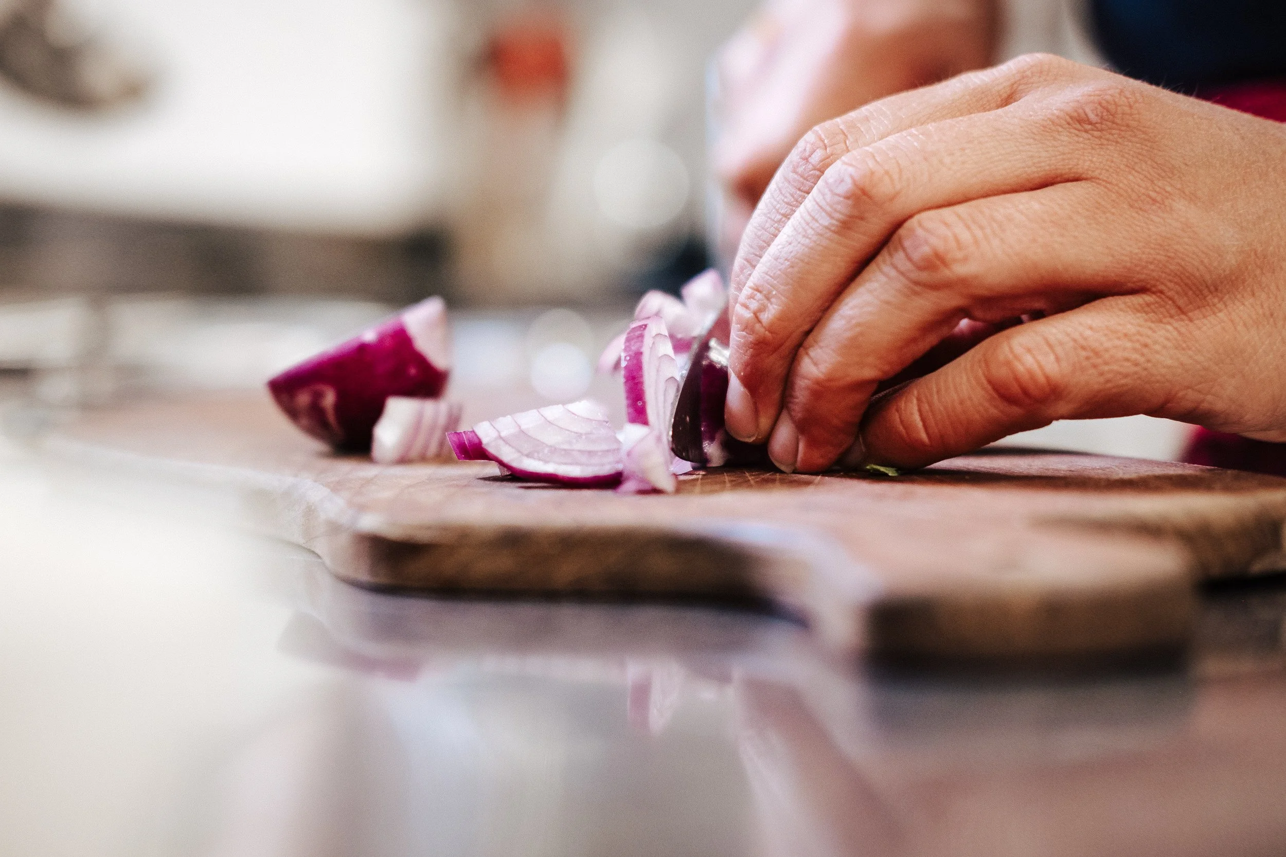 Nahaufnahme einer Hand, die eine Zwiebel auf einem Holzbretthackbrett schneidet, im Hintergrund eine Küche.