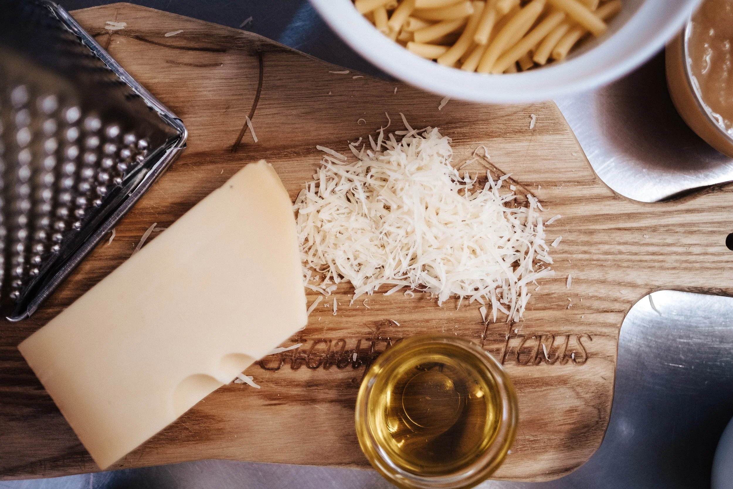 Holzschneidebrett mit geriebenem Käse, einem Stück Käse, einem Glas Öl, Pasta in einer weißen Schüssel, und eine Käsereibe.