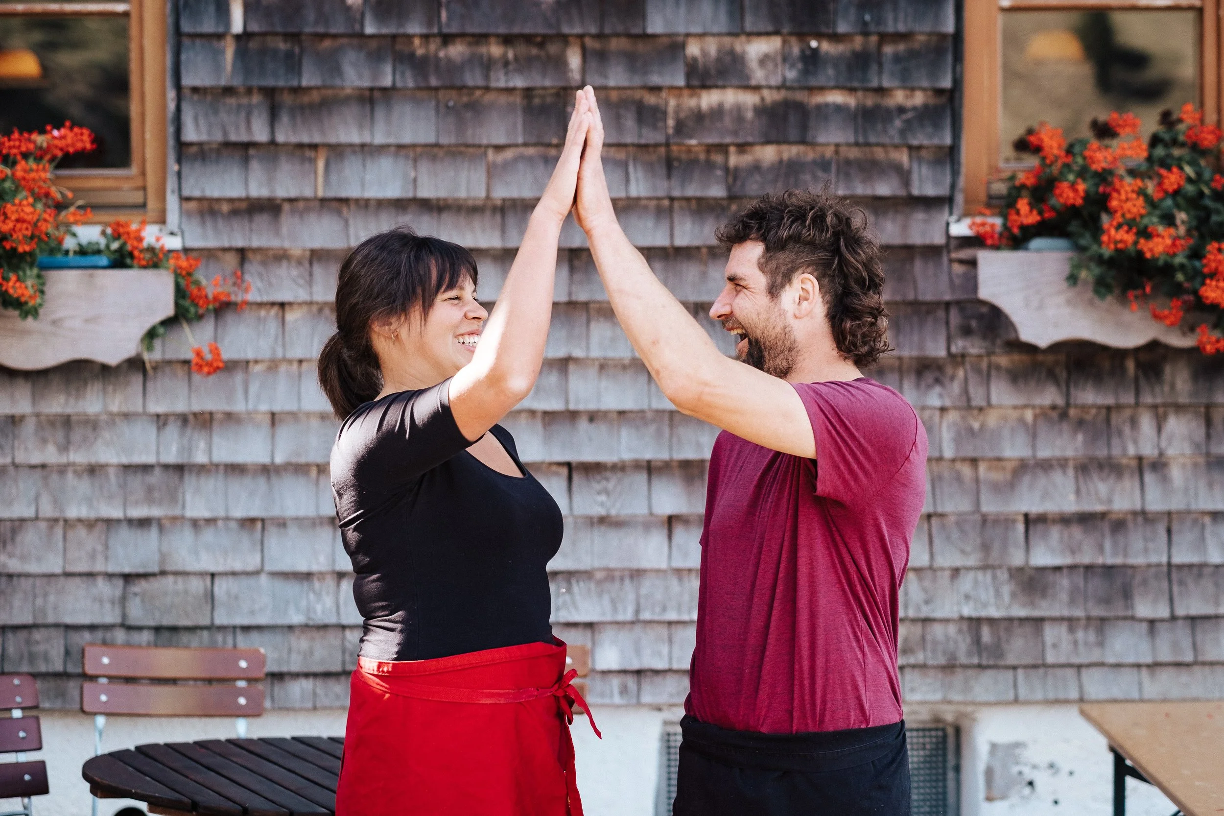 Zwei Menschen, die sich high five geben, vor einem Haus mit Holzwand und Blumenkästen.
