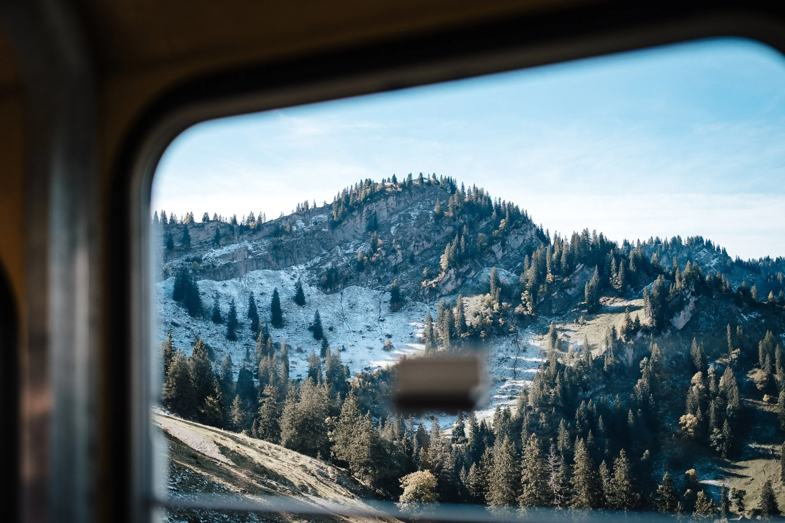 Berglandschaft mit bewaldetem Hügel, gesehen durch ein Fenster eines Zuges oder Fahrzeugs.