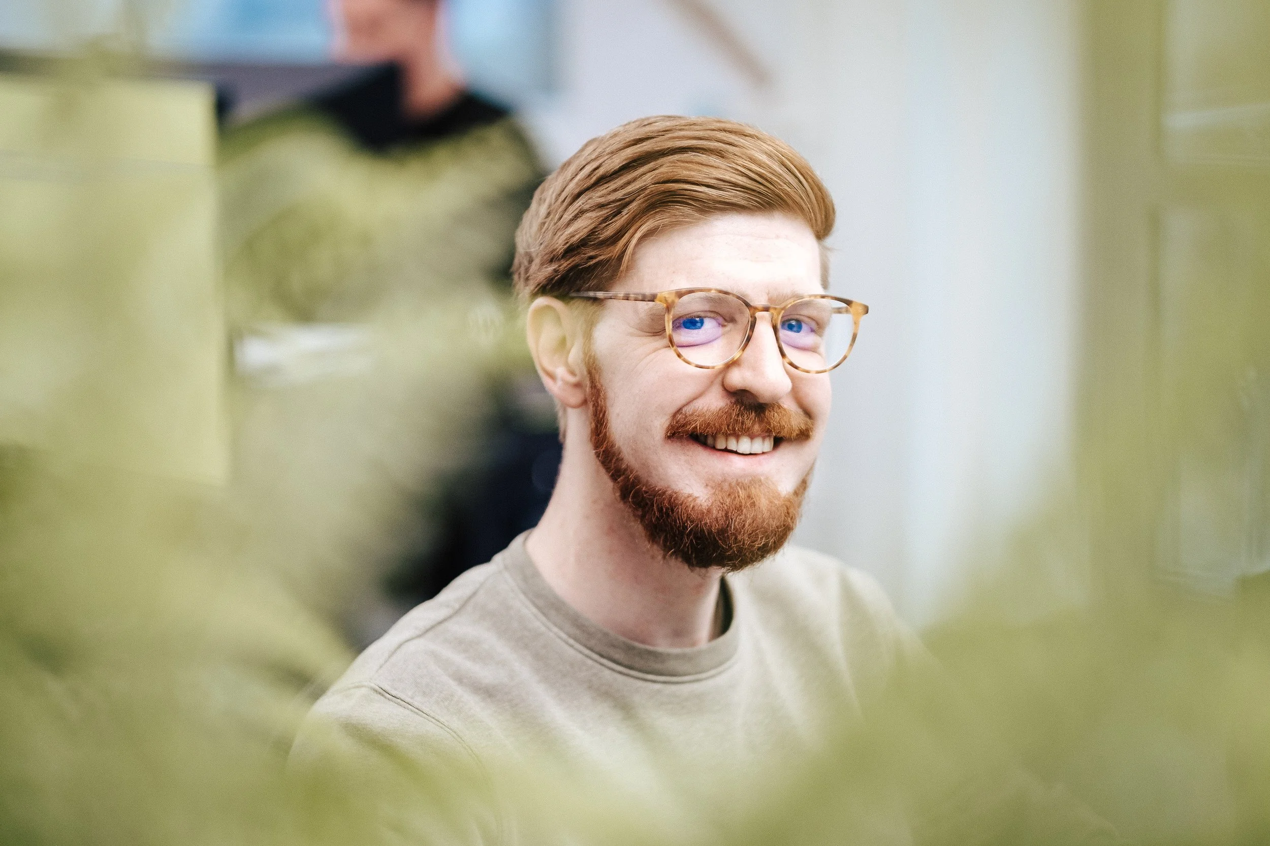 Lächelnder Mann mit Brille und rotem Haar, im Hintergrund unscharf, vorne unscharf grüne Blätter oder Pflanzen, wahrscheinlich in einem Raum oder Büro