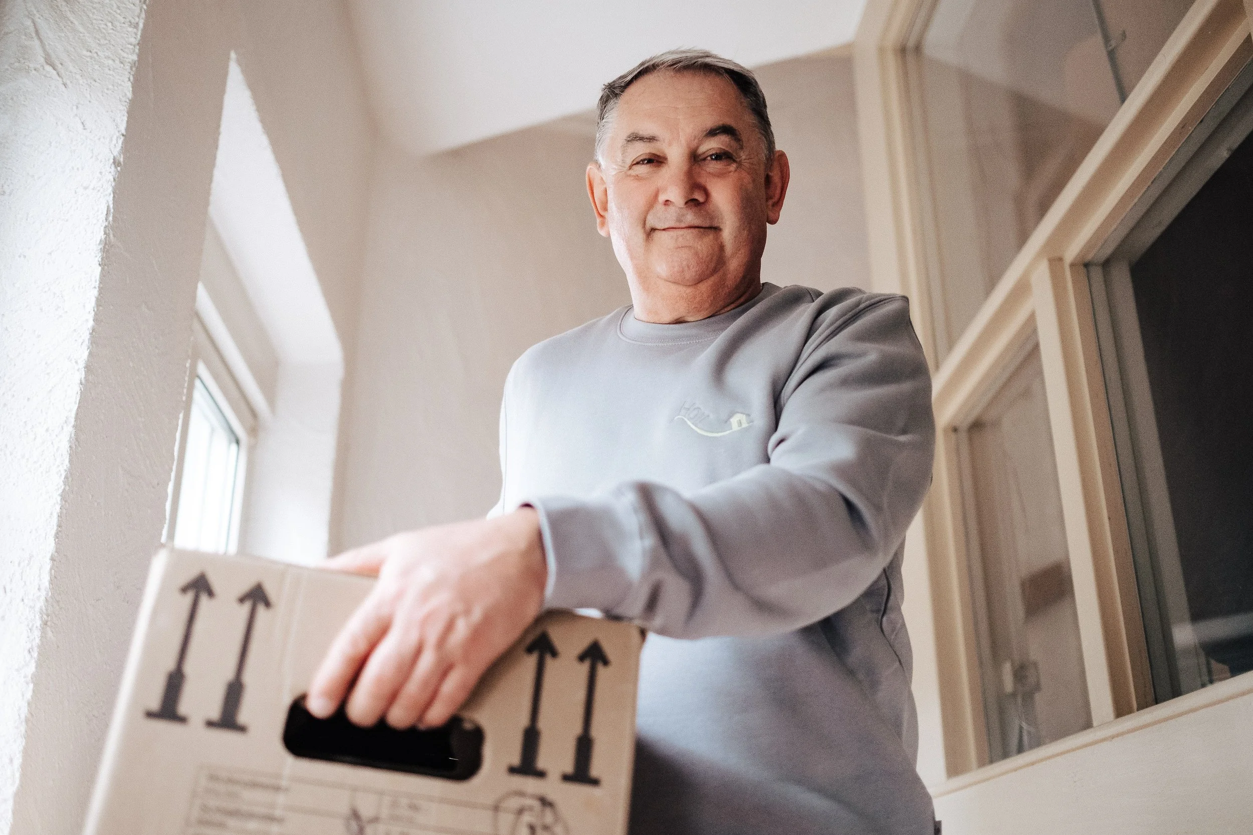 Älterer Mann packt eine Umzugskiste und lächelt in die Kamera in einem hellen Raum.