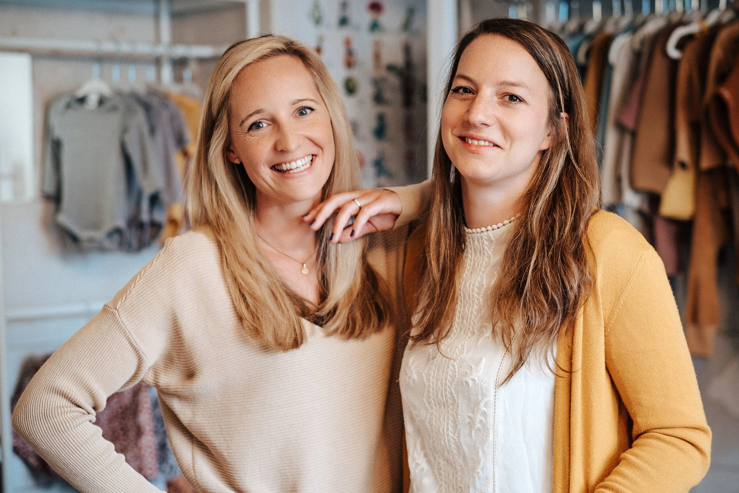 Zwei junge Frauen stehen in einem Kleidungsgeschäft, lächeln und posieren für die Kamera, mit Regalen voller Kleidung im Hintergrund.