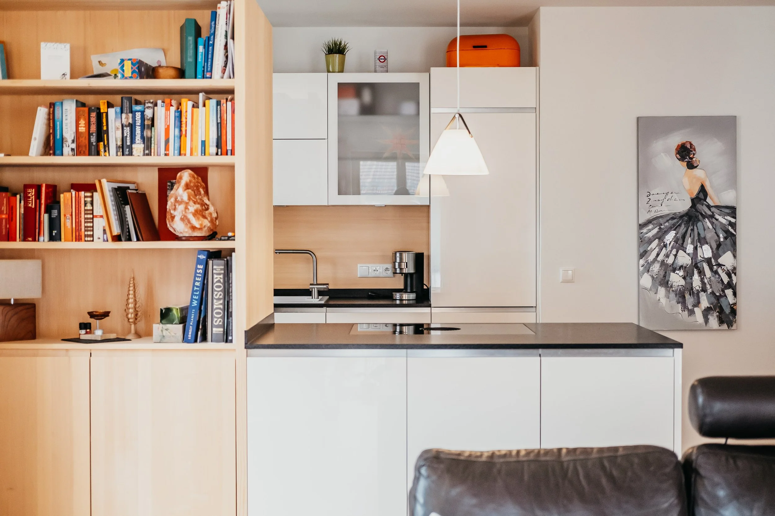 Küche mit weißen Schränken, dunklem Küchenarbeitsplatte, Geschirrspüler, Wasserhahn, Kaffeemaschine, Bücher, Salz- und Pfefferstreuern, Salzlampe, einem Kasten mit Gewürzen, einem Foto der Sonne, einem Glas mit einem warmen Getränk, Kunstwerk eines F