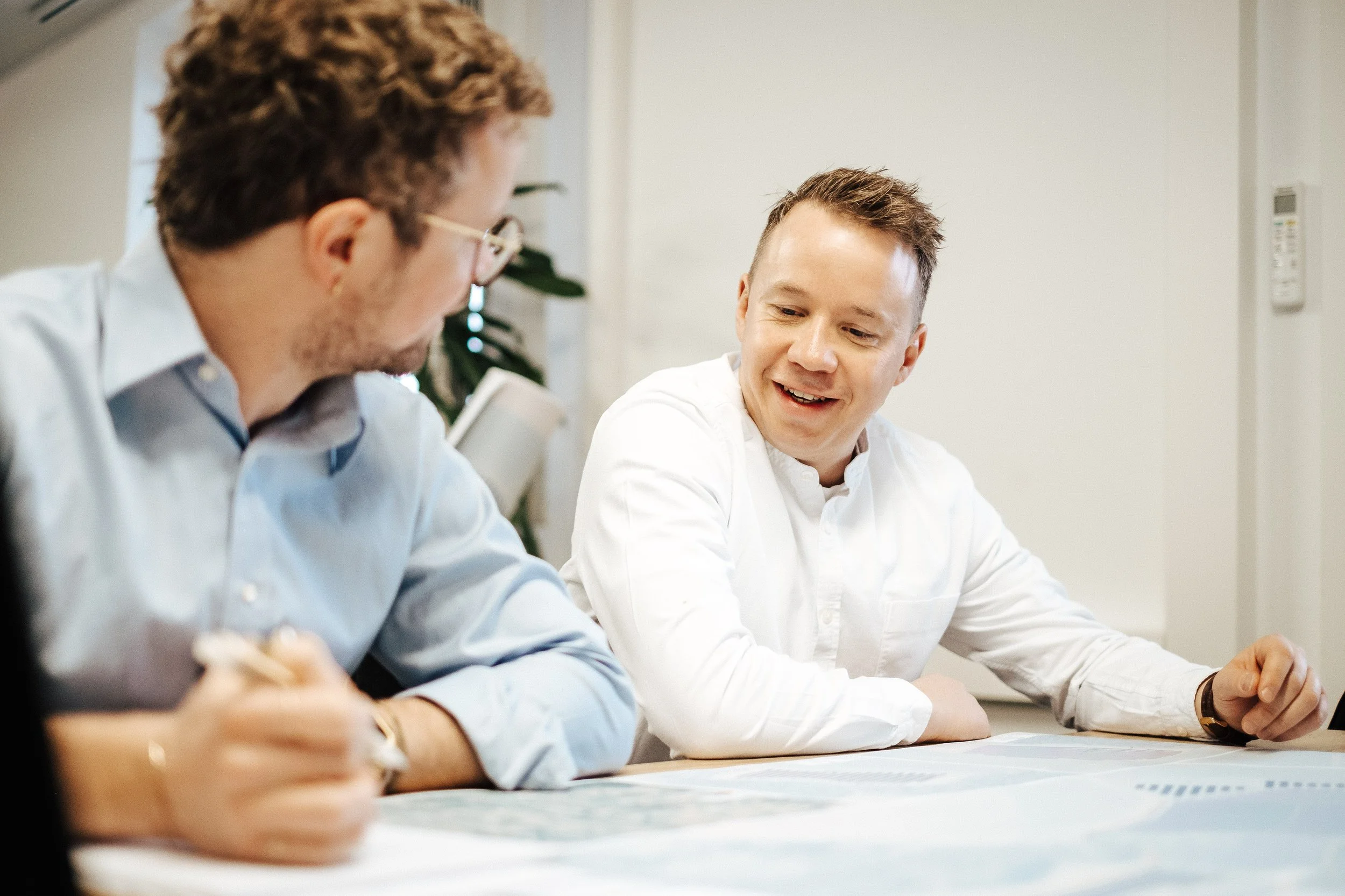 Zwei Männer sitzen an einem Tisch und unterhalten sich, einer schreibt auf Papier, der andere lächelt.