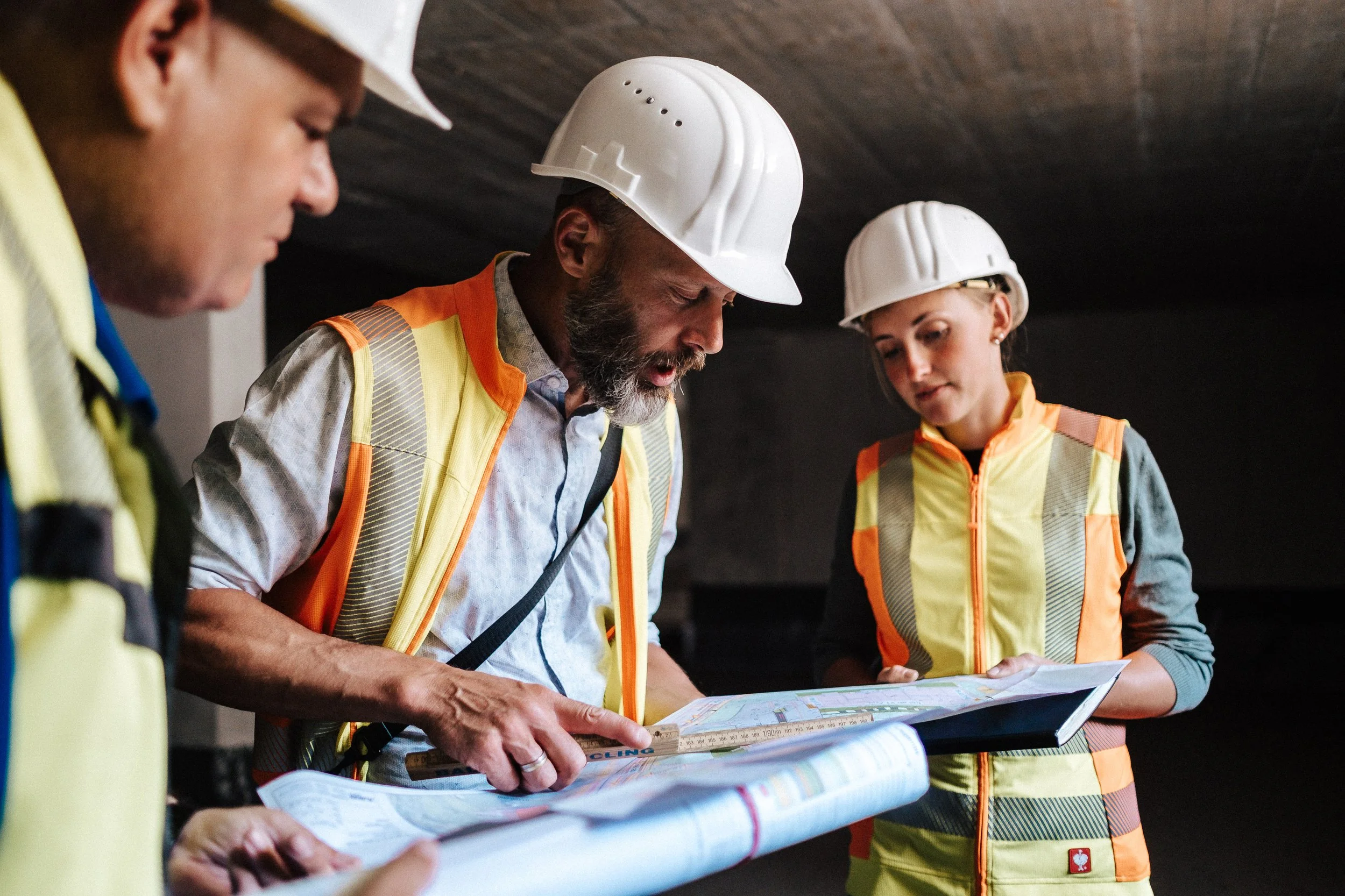 Drei Bauarbeiter in Schutzhelmen und Sicherheitswesten, die im Dunkeln Pläne oder Bauzeichnungen studieren.