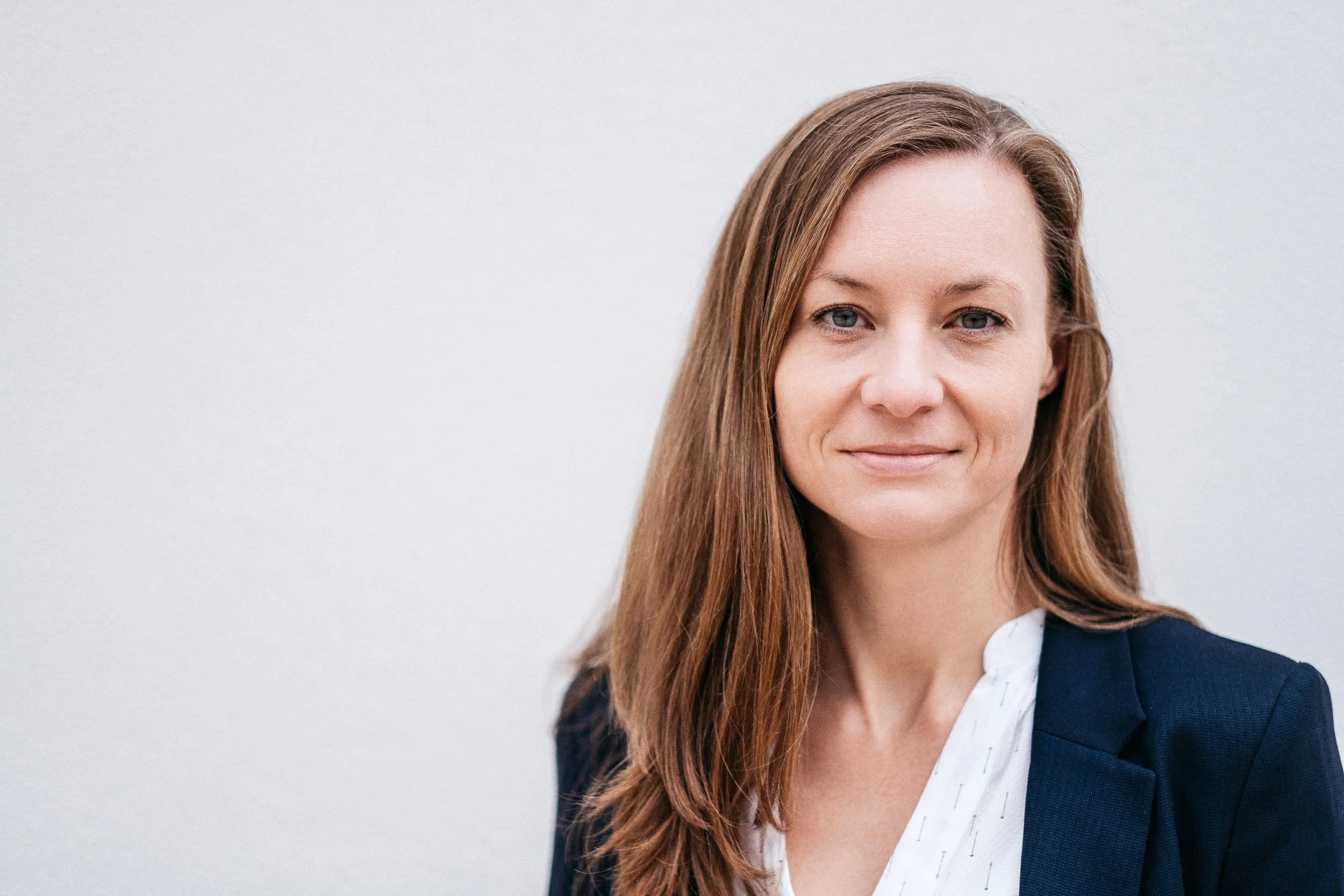 Porträt einer Frau mit braunen Haaren, hellem Teint, blauen Augen, in dunklem Blazer und weißer Bluse vor neutralem Hintergrund.