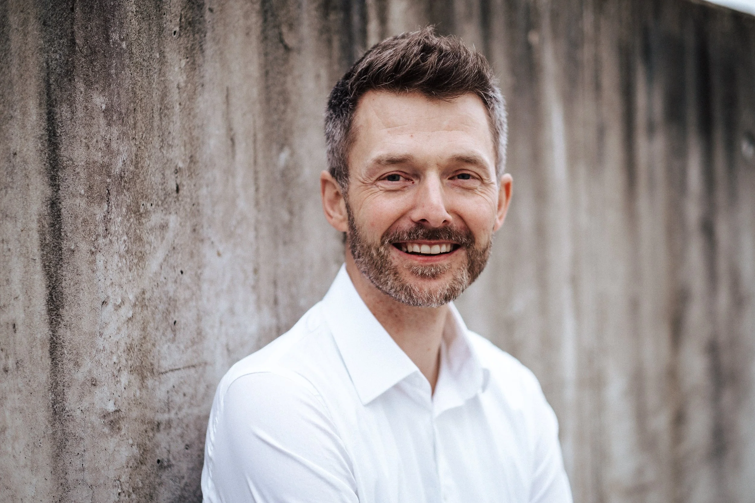 Lächelnder Mann in weißem Hemd steht an einer Betonmauer im Freien.
