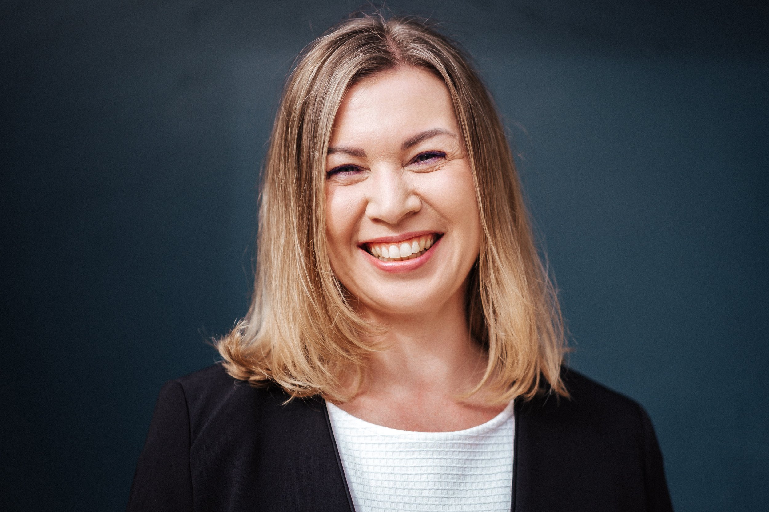 Lächelnde Frau mit blonden Haaren, wearing eine schwarze Jacke und weißen top, unterm Hintergrund ist dunkelblau.