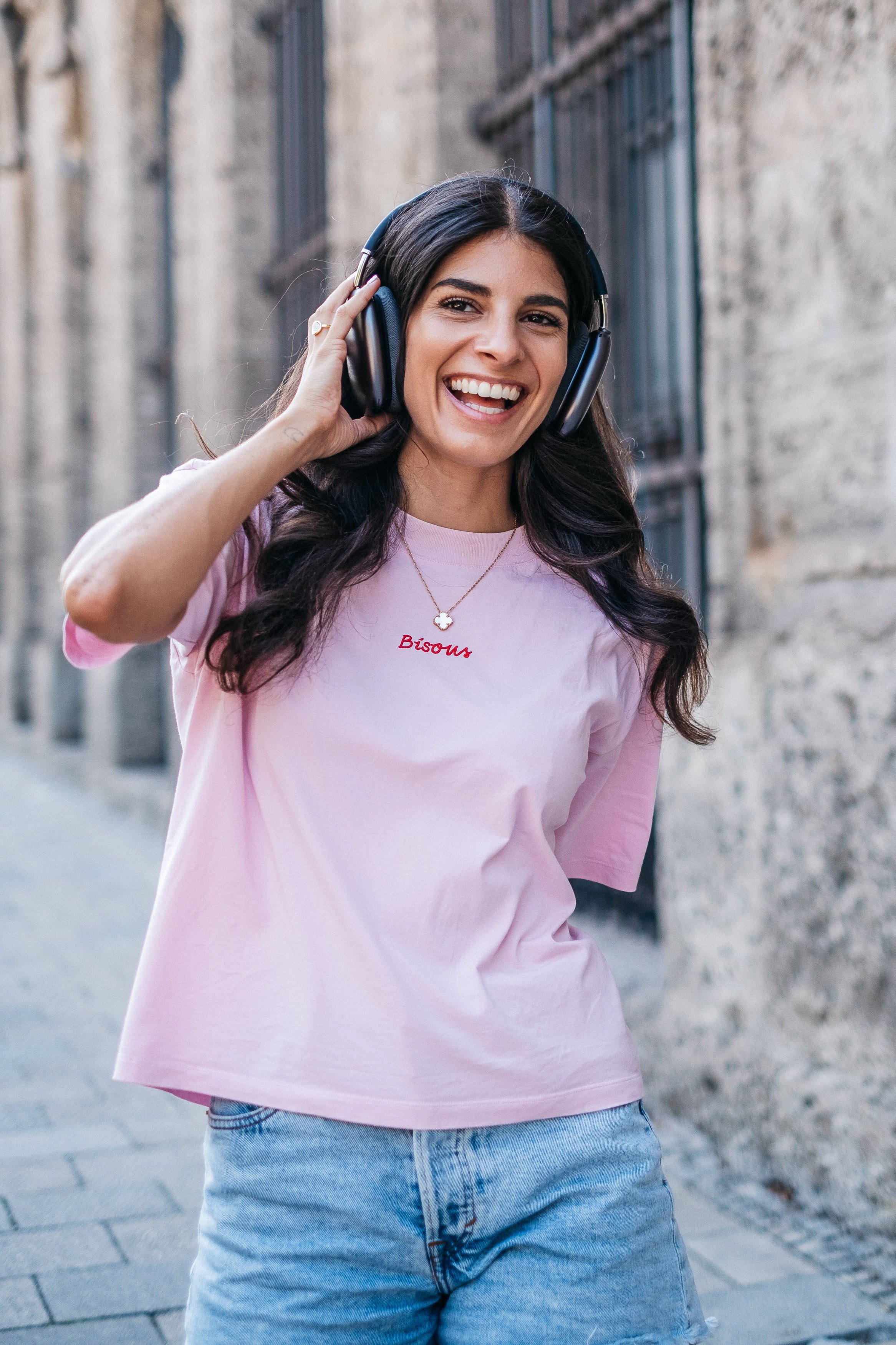 Eine junge Frau trägt Kopfhörer, lächelt und steht in einer Stadtstraße, trägt ein rosa T-Shirt mit der Aufschrift 'Bisous'.