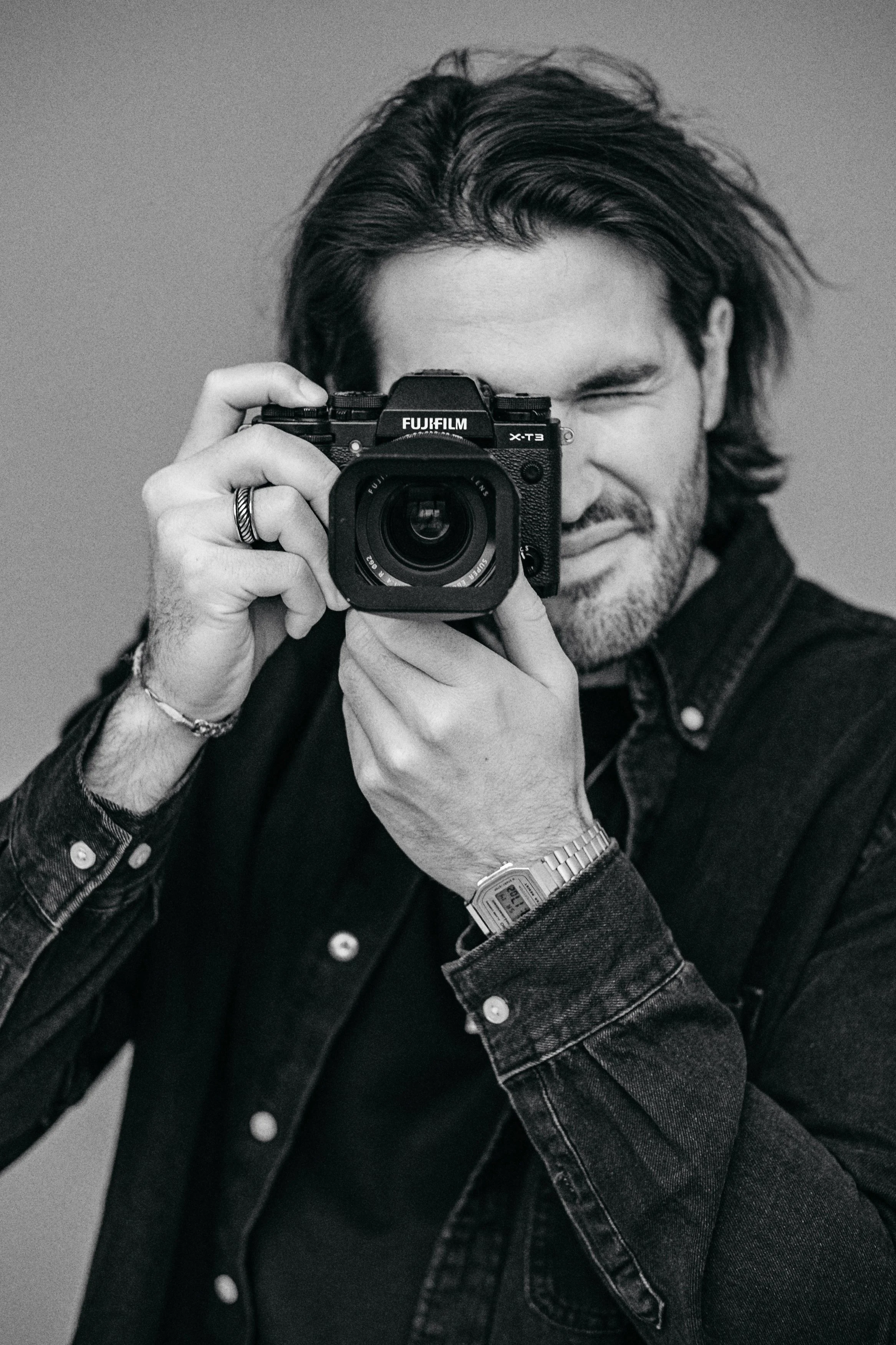 Schwarz-weiß Portrait eines jungen Mannes mit Kamera, trägt dunklen Hemd und Uhr, blickt durch Kamera.