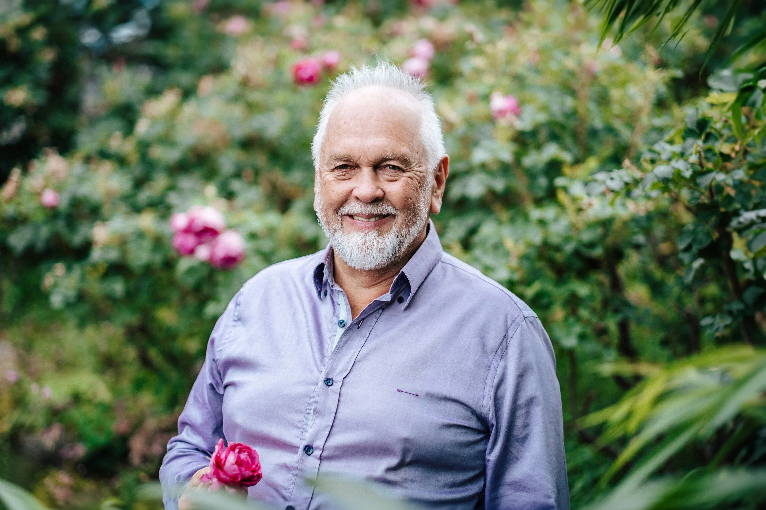 Ein älterer Mann mit weißem Bart und grauem Haar, lächelnd, in einem Garten mit rosa Blumen im Hintergrund.
