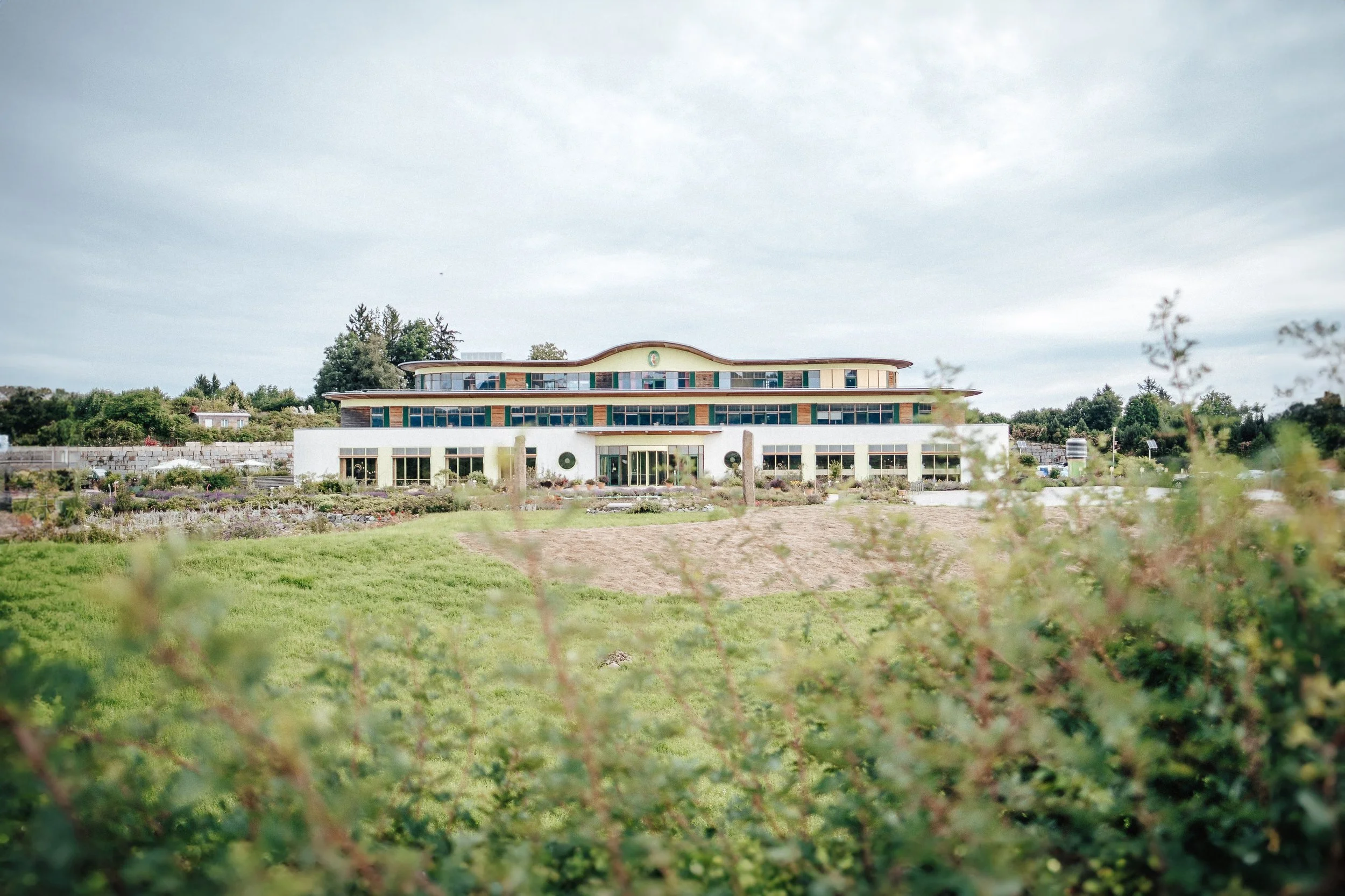 Ein modernes, mehrstöckiges Gebäude inmitten einer grünen Landschaft mit Pflanzen und Feldern, unter wolkigem Himmel.