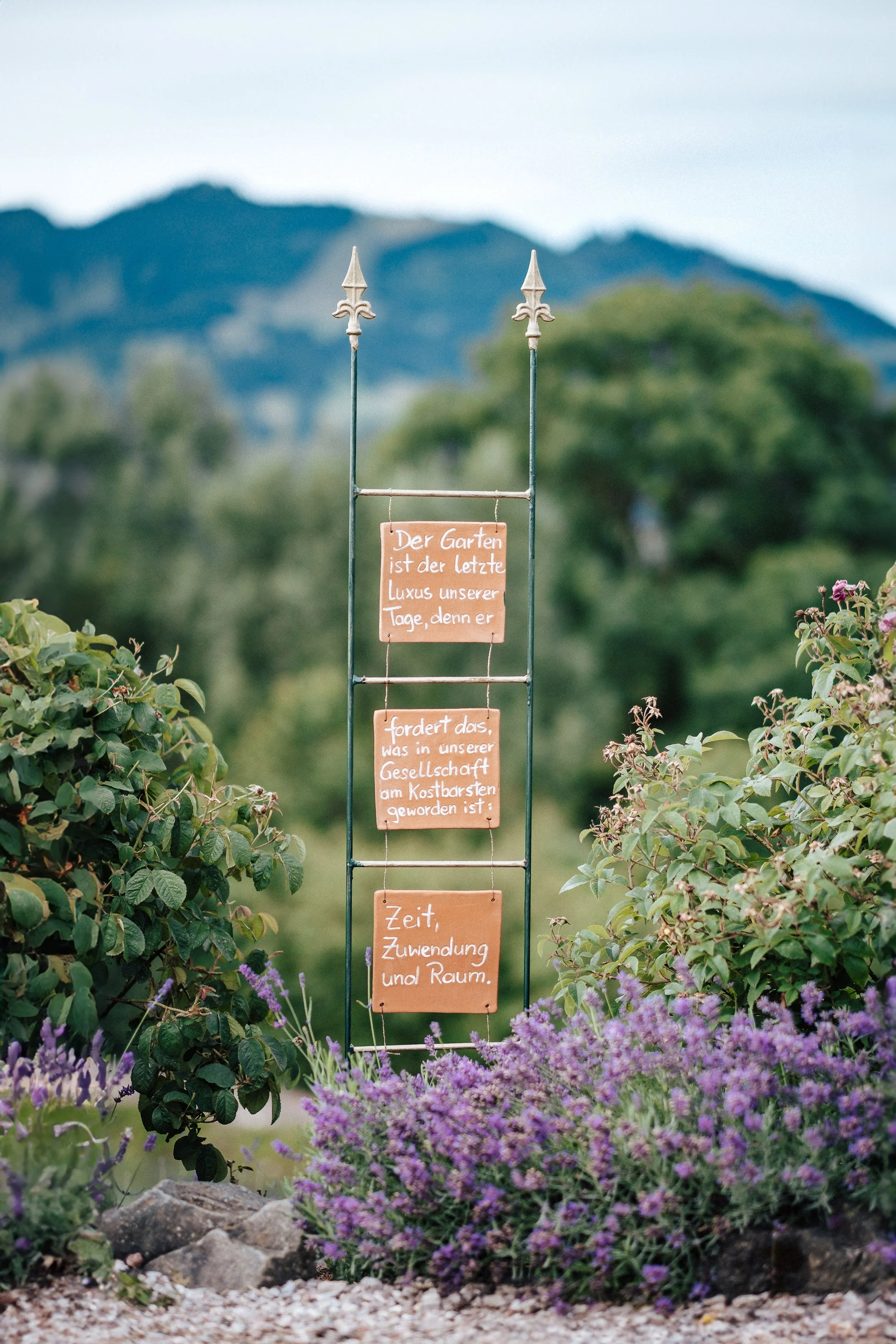 Ein Garten mit einer Metalltafel, auf der ein deutschs Text steht. Der Text lautet: "Der Garten ist der letzte Luxus unserer Tage, denn er fordert das, was in unserer Gesellschaft am kostbarsten geworden ist - Zeit, Zuwendung und Raum." Der Garten is