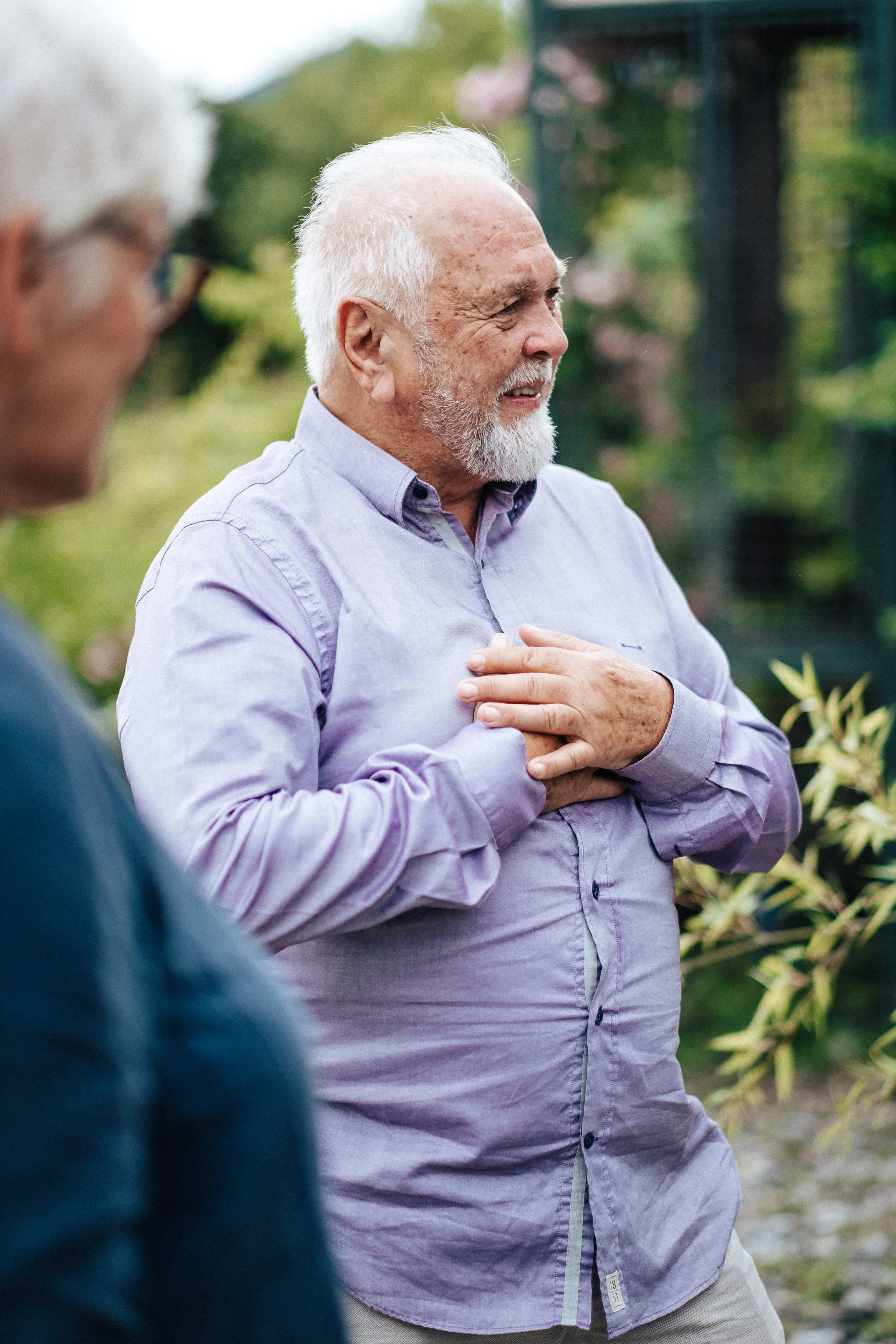 Ein älterer Mann mit grauem Bart, der eine Hand auf die Brust gelegt hat, während er spricht oder singt, im Freien bei schönem Wetter.