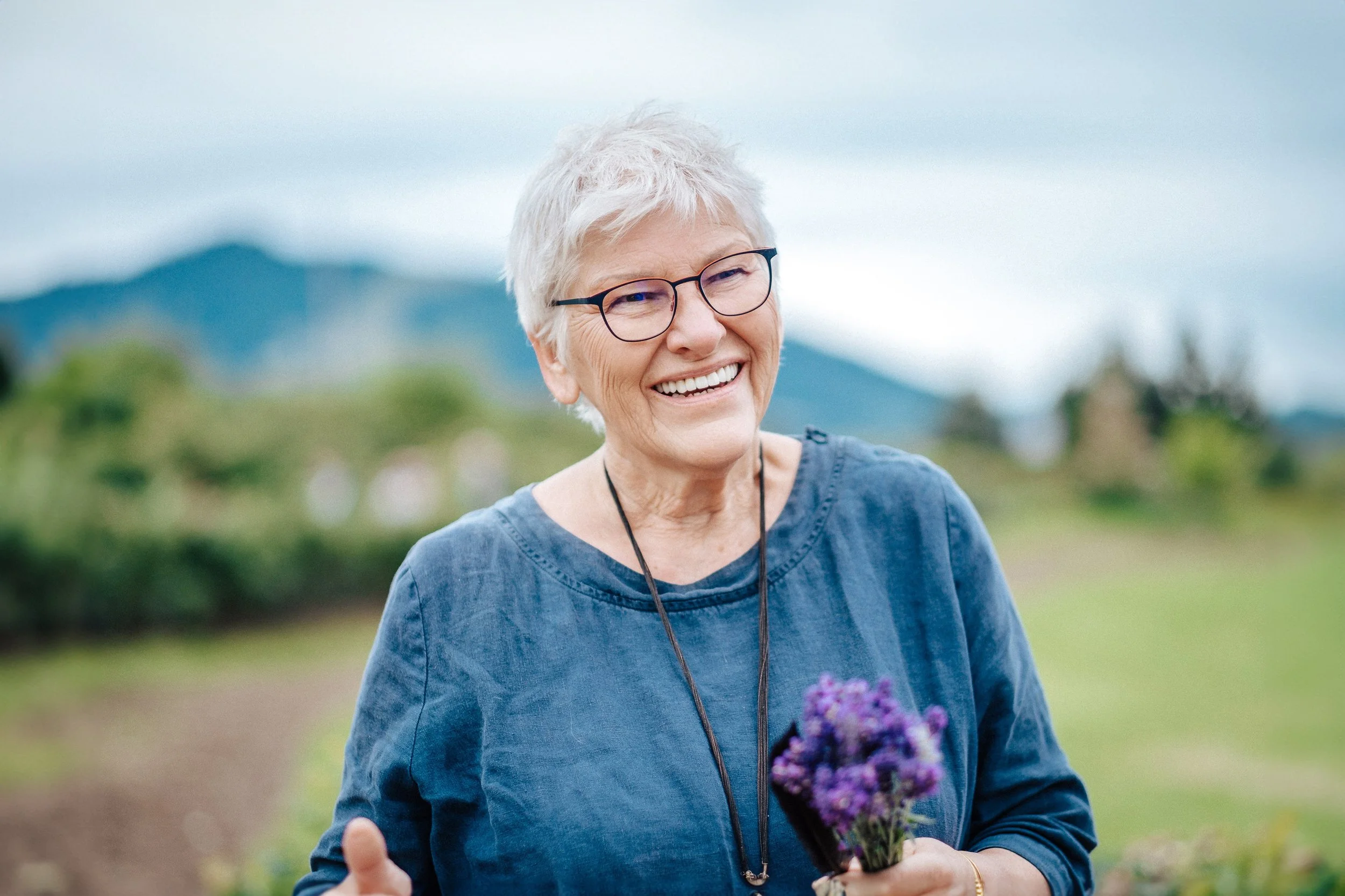 Eine ältere Frau mit kurzen grauen Haaren, Brille und dunklem Shirt lächelt und hält einen kleinen Blumenstrauß in der Hand, im Hintergrund Berge und Natur.