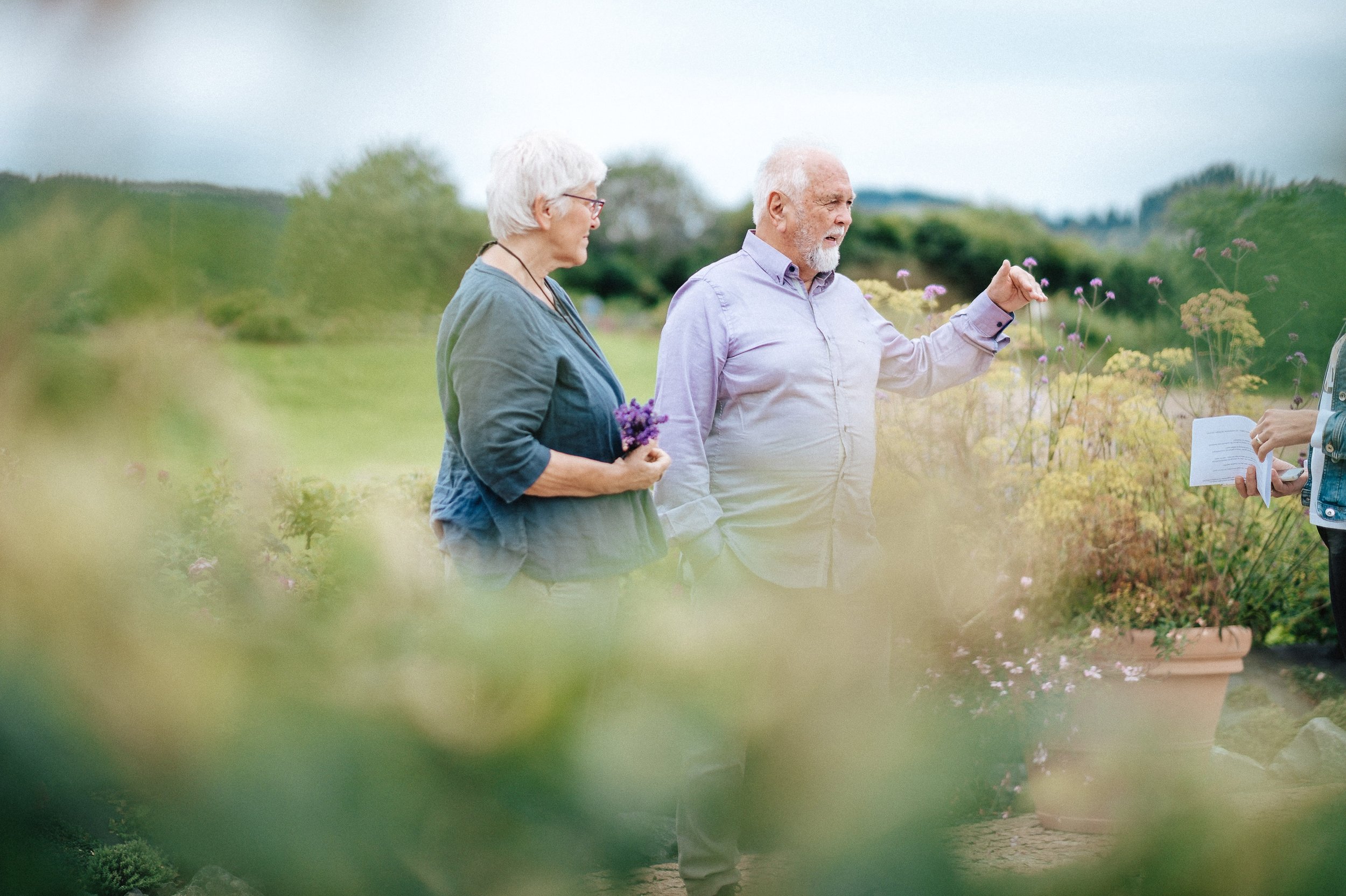 Zwei ältere Menschen, ein Mann und eine Frau, stehen in einem Garten oder Park und unterhalten sich. Sie sind umgeben von bunten Blumen und Pflanzen, während im Hintergrund eine grüne Landschaft und ein Himmel mit Wolken sichtbar sind.