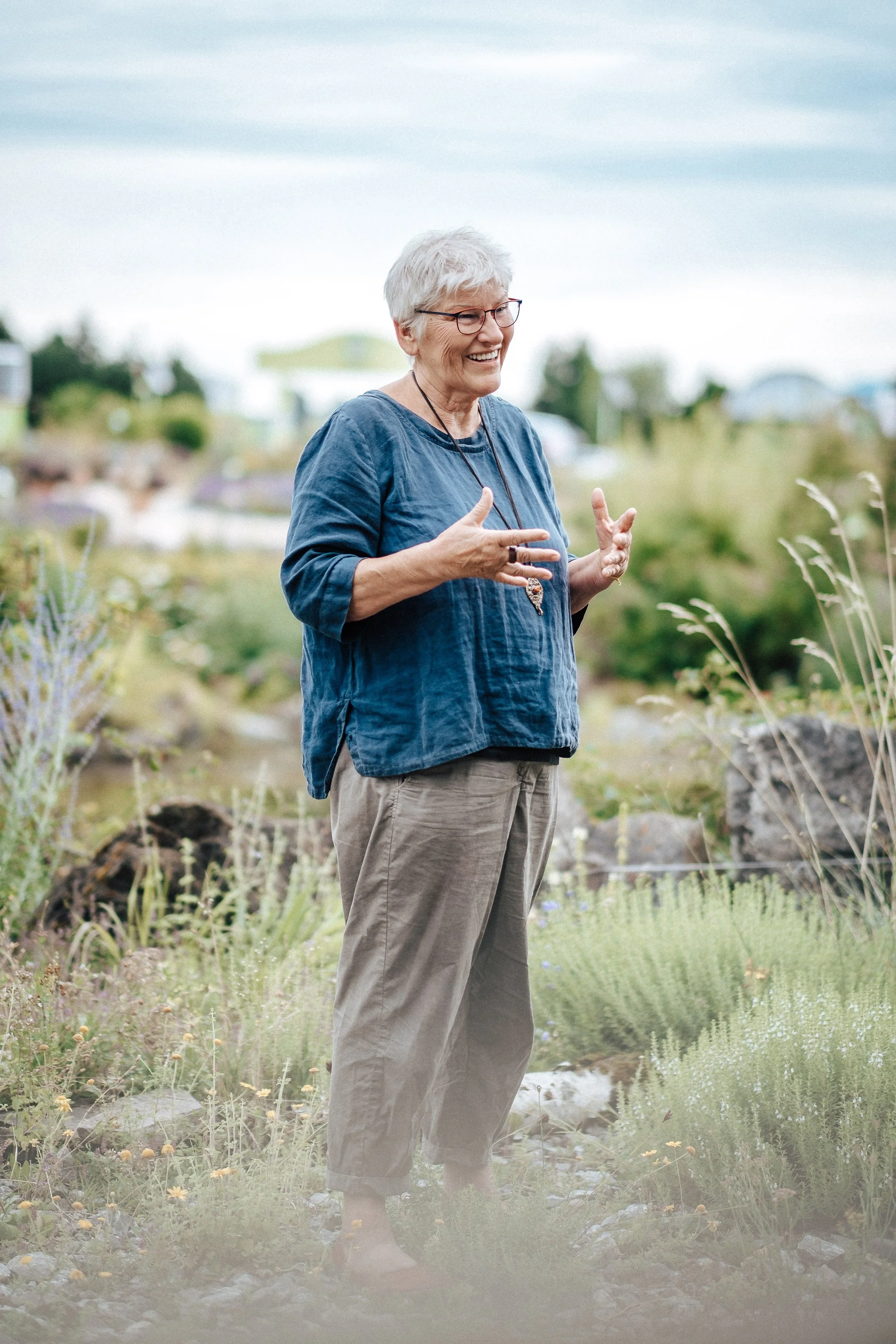 Eine ältere Frau mit kurzen grauen Haaren, trägt eine blaue Bluse und beige Hosen, steht im Freien in einer Gartenlandschaft und lacht, während sie spricht.