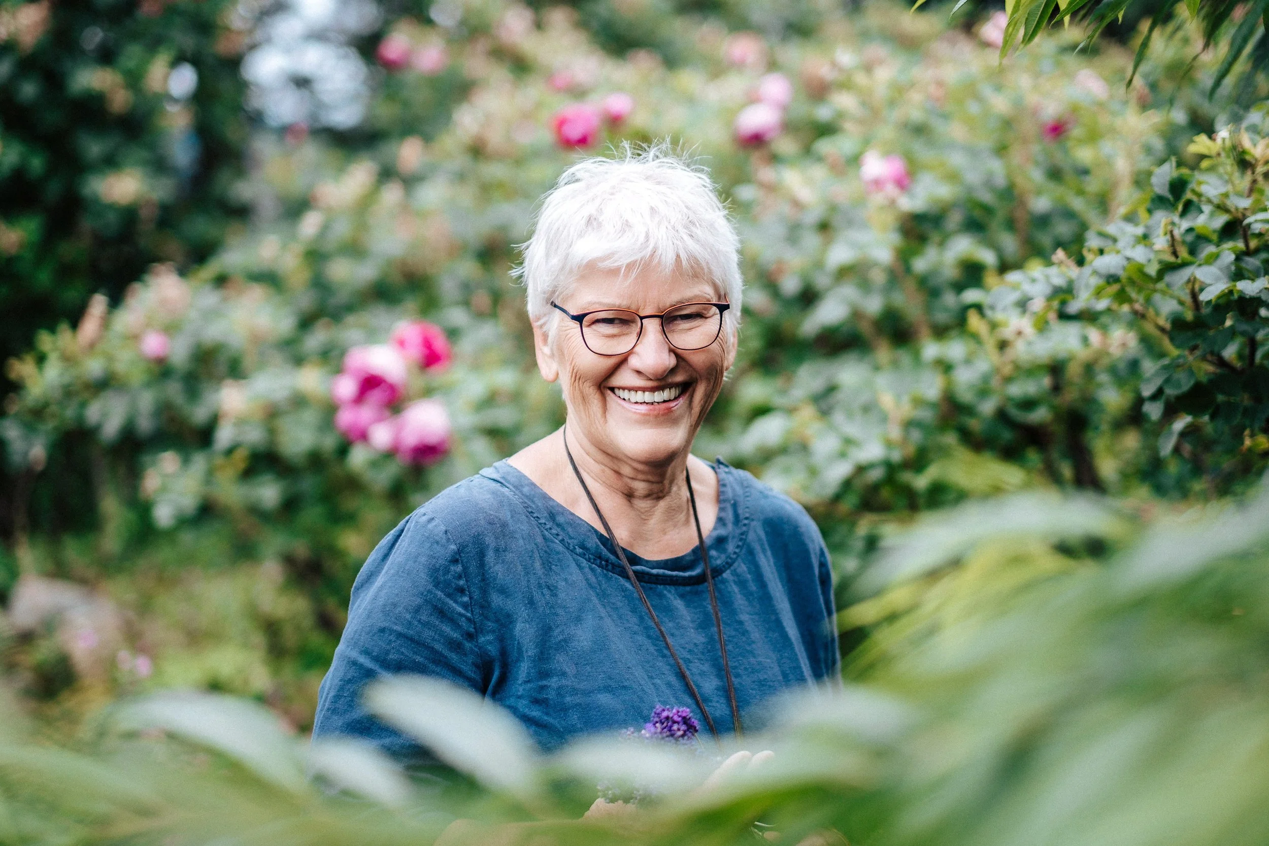 Lächelnde ältere Frau mit weißen Haaren und Brille in einem Garten mit blühenden Rosen, trägt eine blaue Bluse.