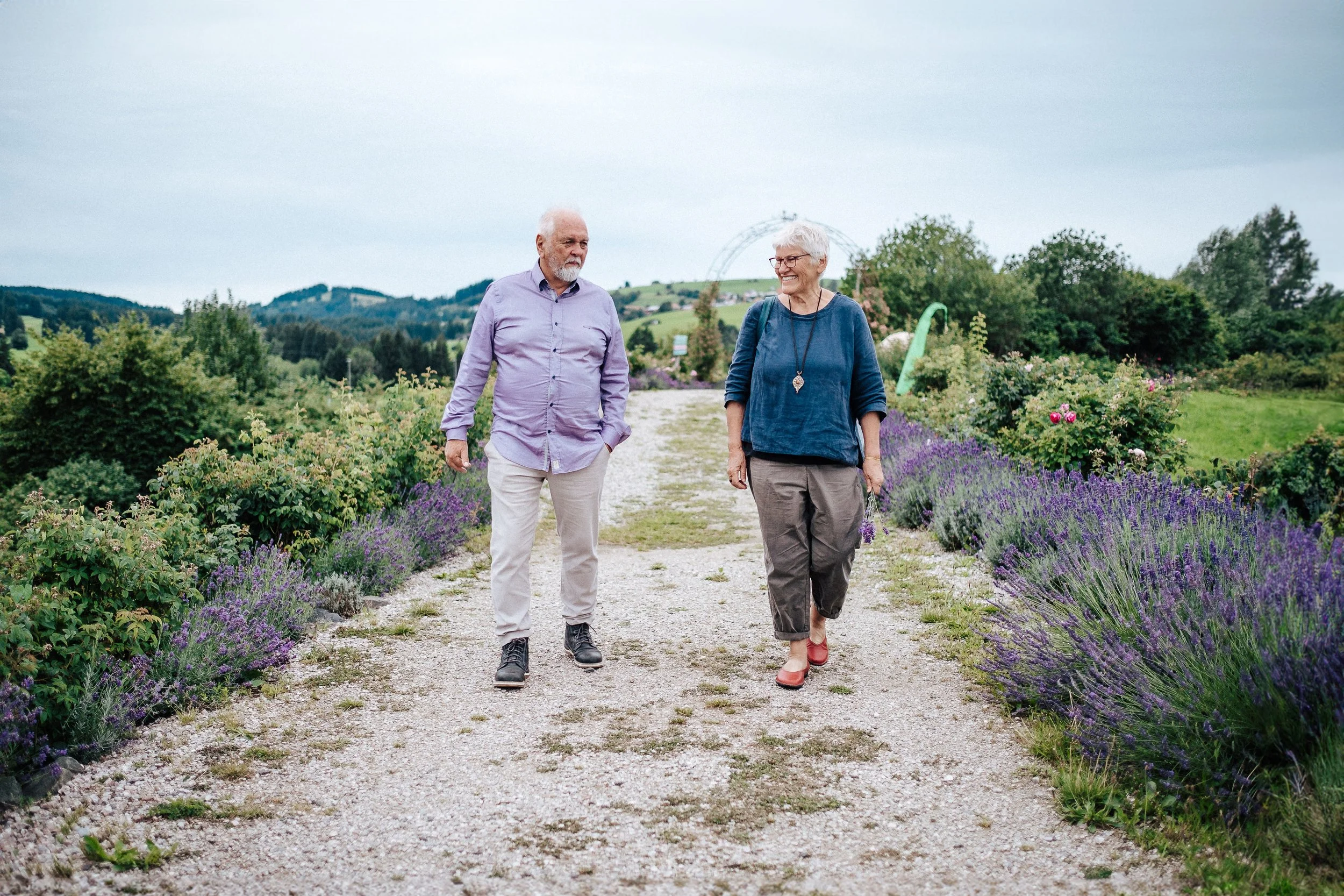 Ein älteres Paar spaziert auf einem Weg in einem Lavendelfeld mit grünen Hügeln im Hintergrund, bei bewölktem Himmel.