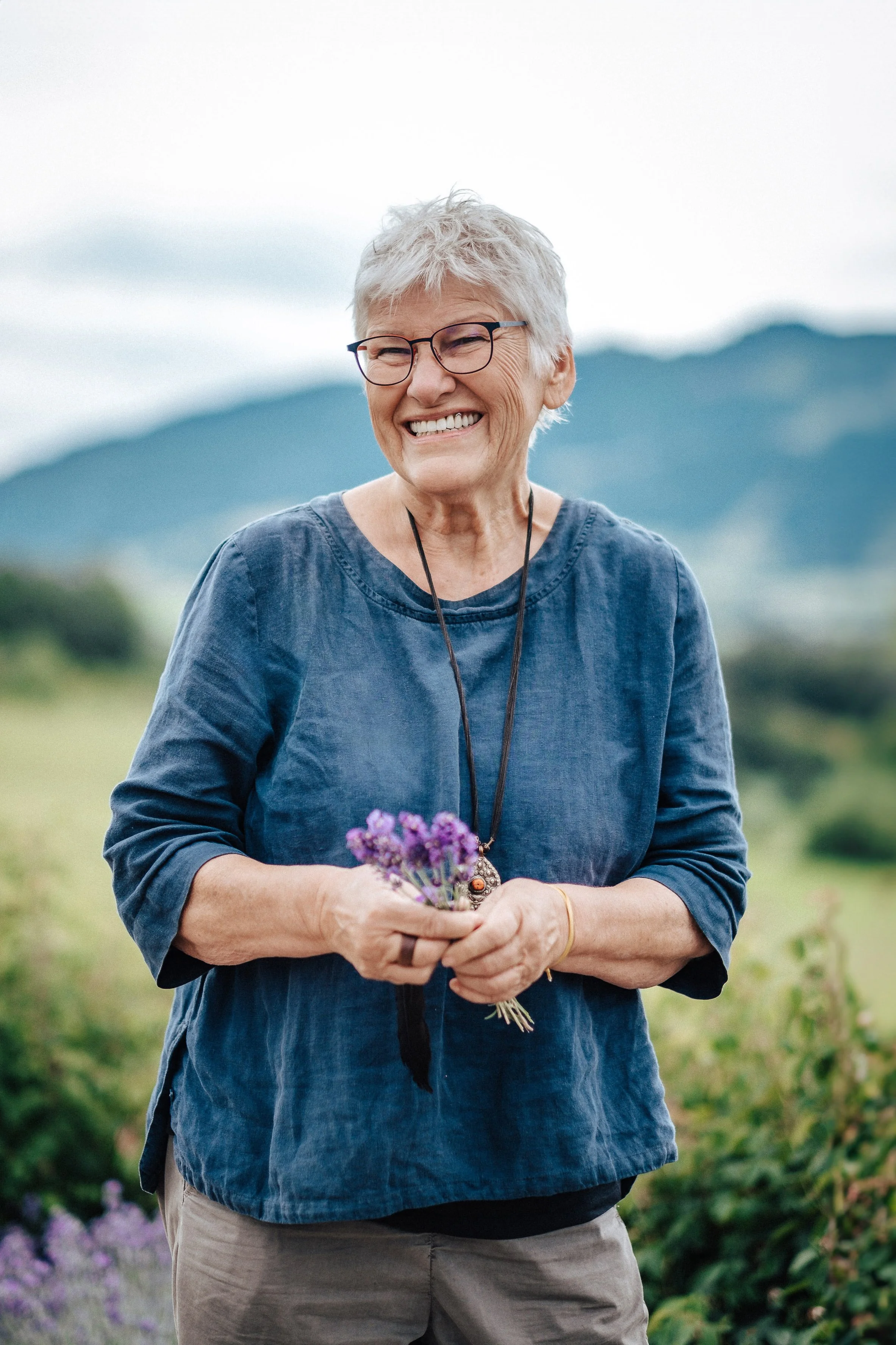 Lächelnde ältere Frau mit Brille steht im Freien, hält einen kleinen Blumenstrauß aus Lavendel, im Hintergrund Berge und eine grüne Landschaft.