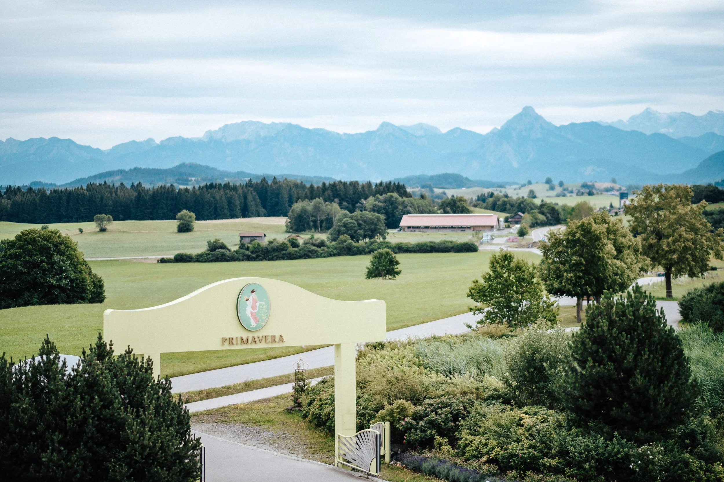 Landschaft mit grünen Wiesen, Bäumen und Bergen im Hintergrund, ein Eingangsschild mit dem Wort 'Primavera' und einem Logo, das eine nackte Frau in einem Kleid zeigt.
