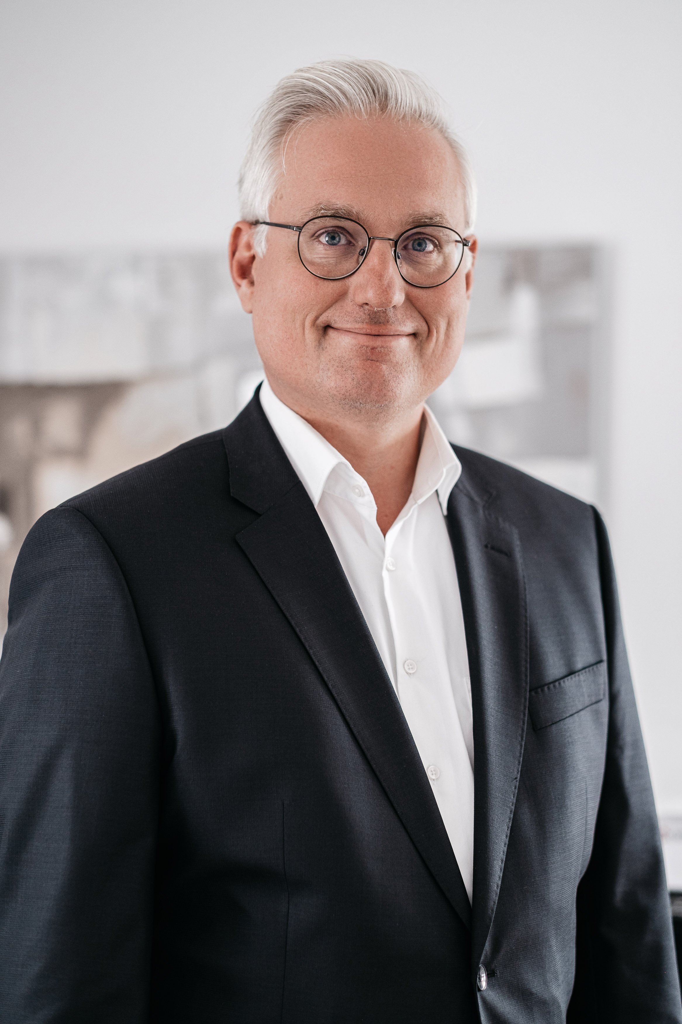 Ein älterer Mann mit grau Haaren, Brille und einem schwarzen Anzug, der in einem Büro steht und lächelt.