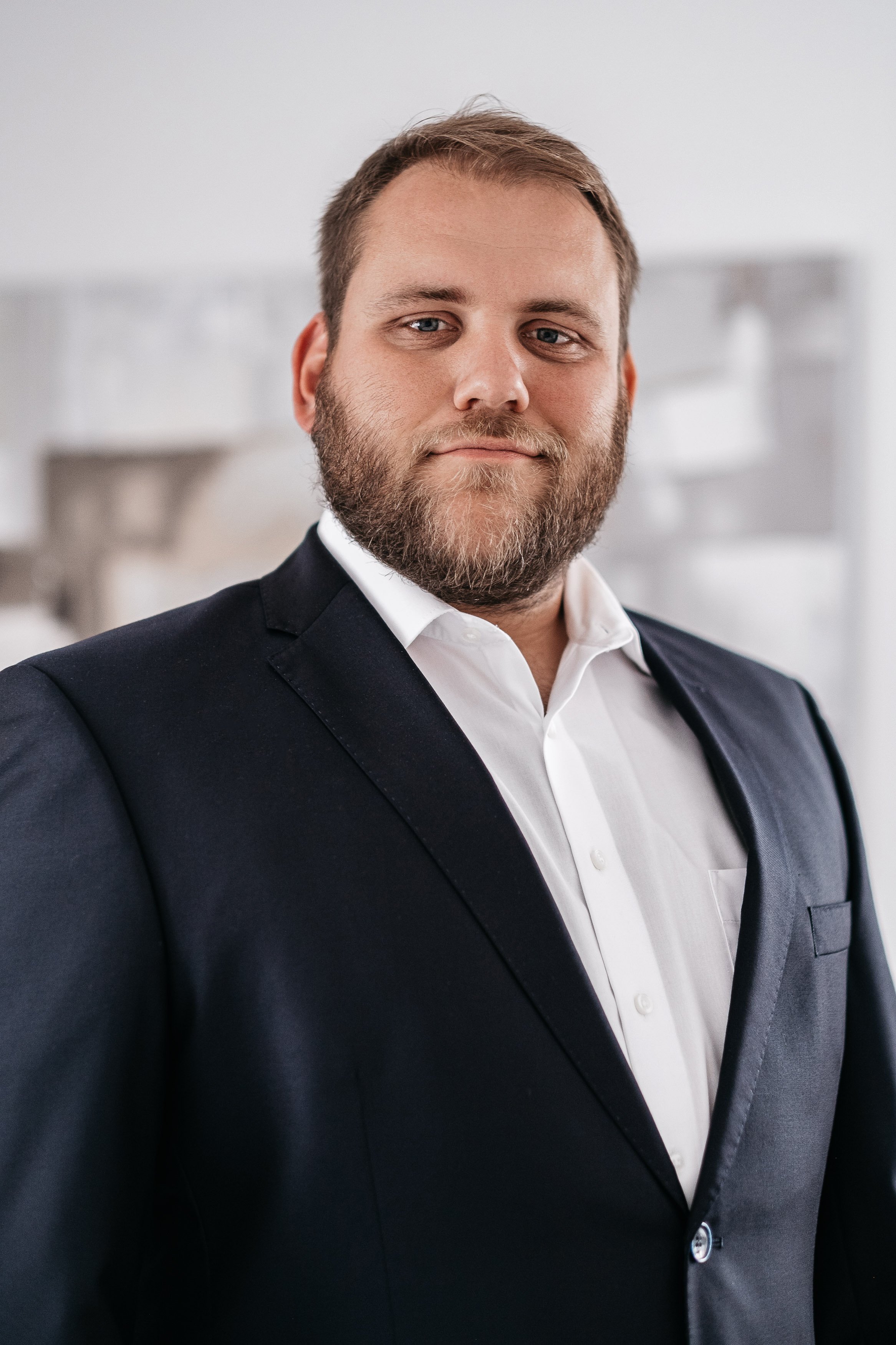 Ein Mann trägt einen dunklen Anzug und ein weißes Hemd, steht vor einem neutralen Hintergrund, lächelt leicht und schaut in die Kamera.