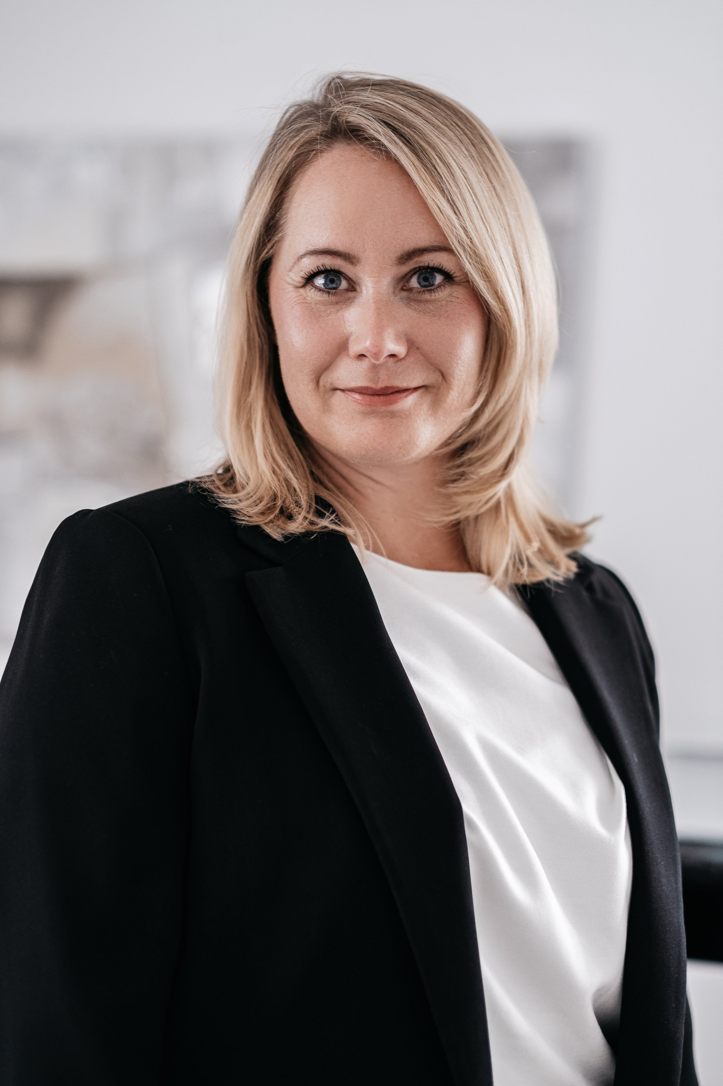 Porträt einer lächelnden Frau mit blonden, kinnlangen Haaren, in einem schwarzen Blazer und weißem Oberteil, vor einem grauen Hintergrund.