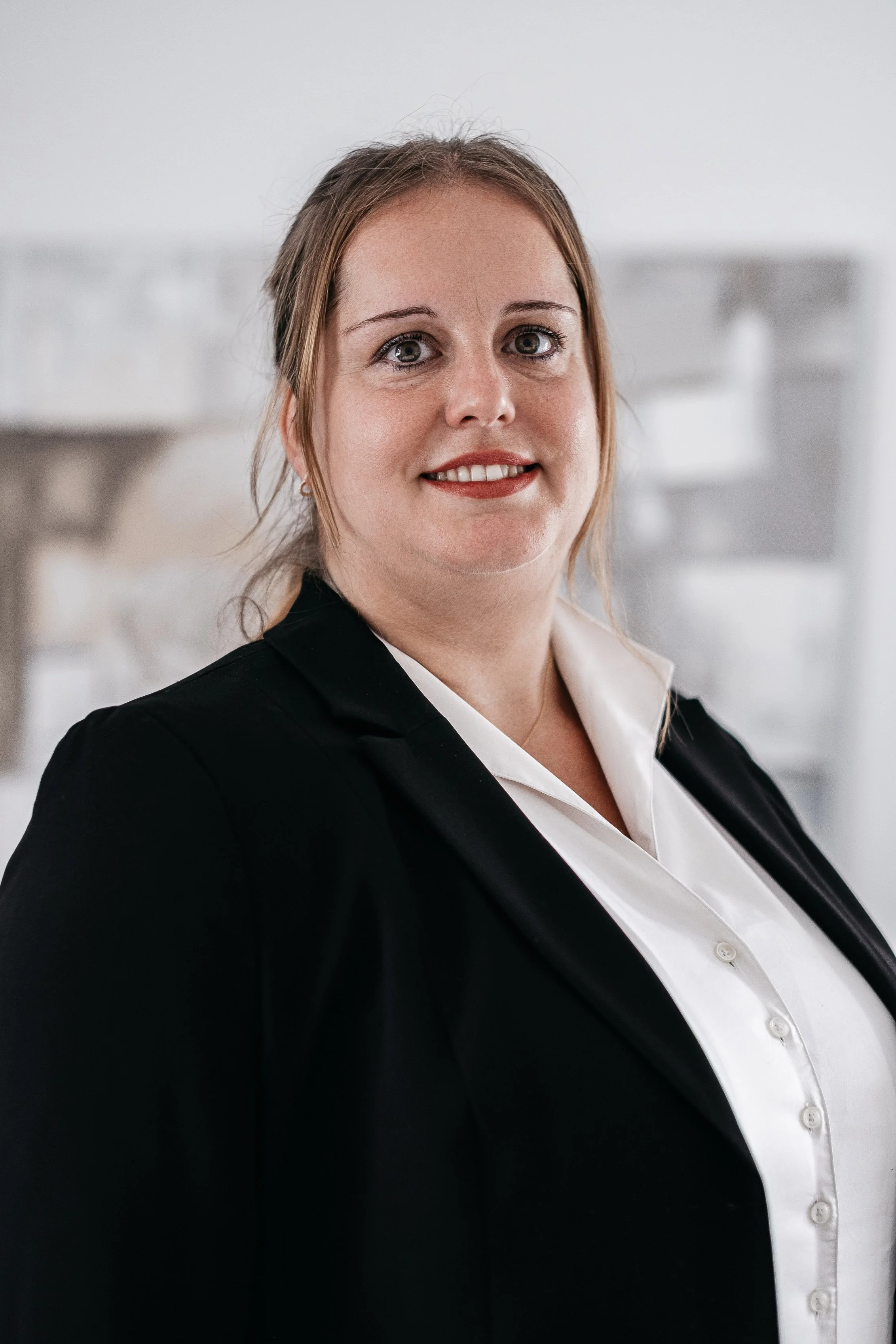 Porträt einer Frau in formeller Kleidung, schwarze Blazer und weiße Bluse, vor hellem, verschwommenem Hintergrund.