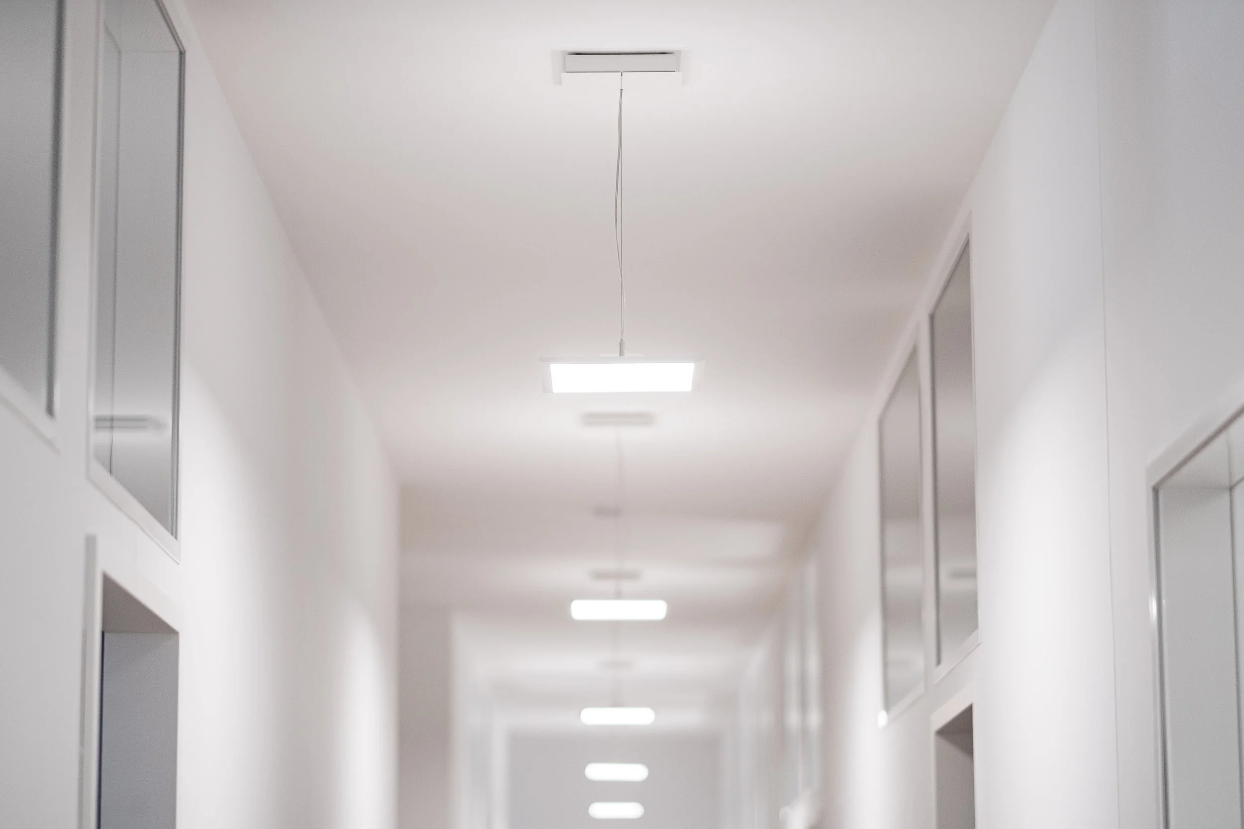 Weißer Flur mit Deckenleuchten und Fenstern auf beiden Seiten, Blick in die Tiefe.