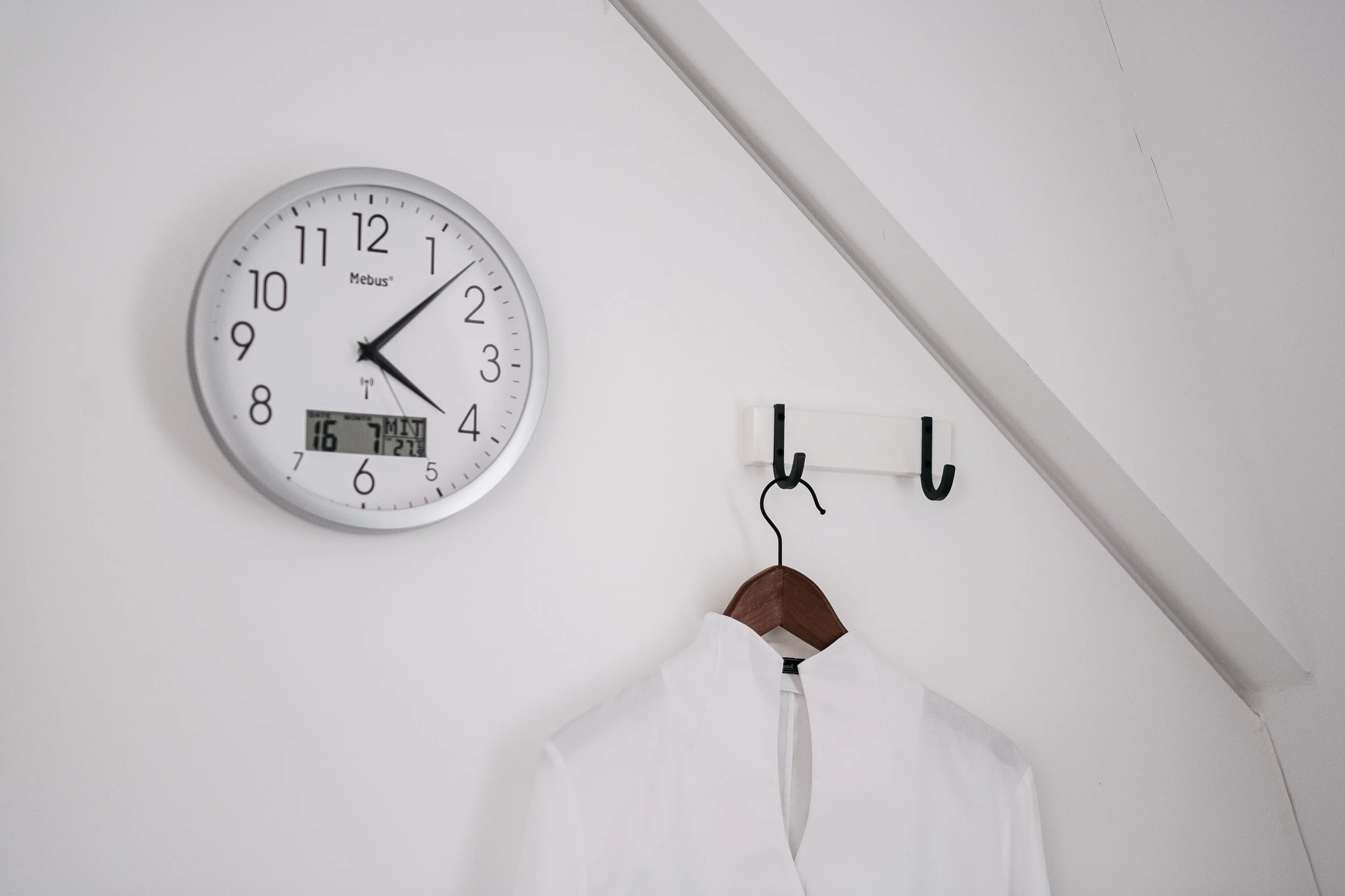 Weiße Wand mit Wanduhr, die die Uhrzeit 13:21 zeigt, daneben eine Kleiderhakenleiste mit einem weißen Hemd und einem schwarzen Haken.