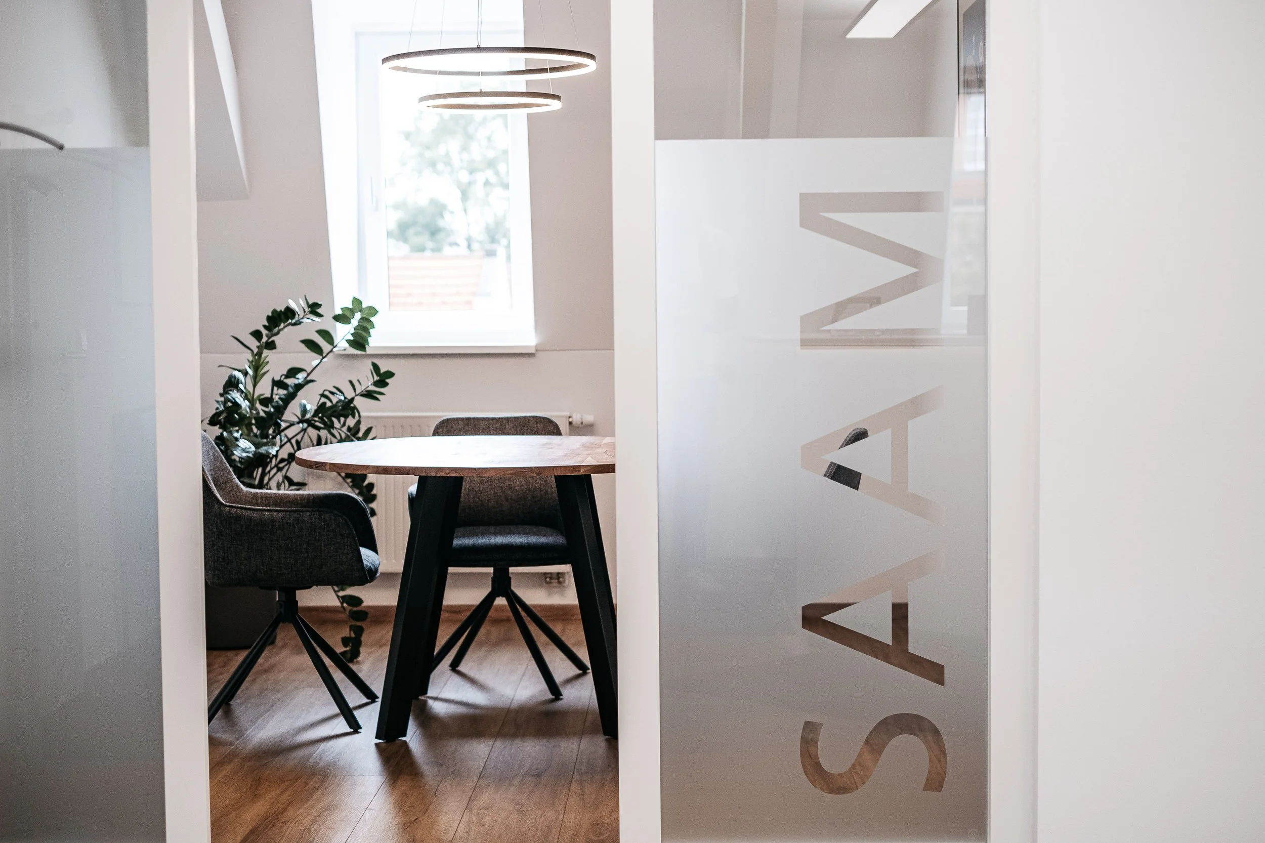 Ein Büroraum mit einem runden Tisch, drei dunkle Stühle und einer Pflanze durch ein beschriftetes Glaselement mit dem Wort "SAY"