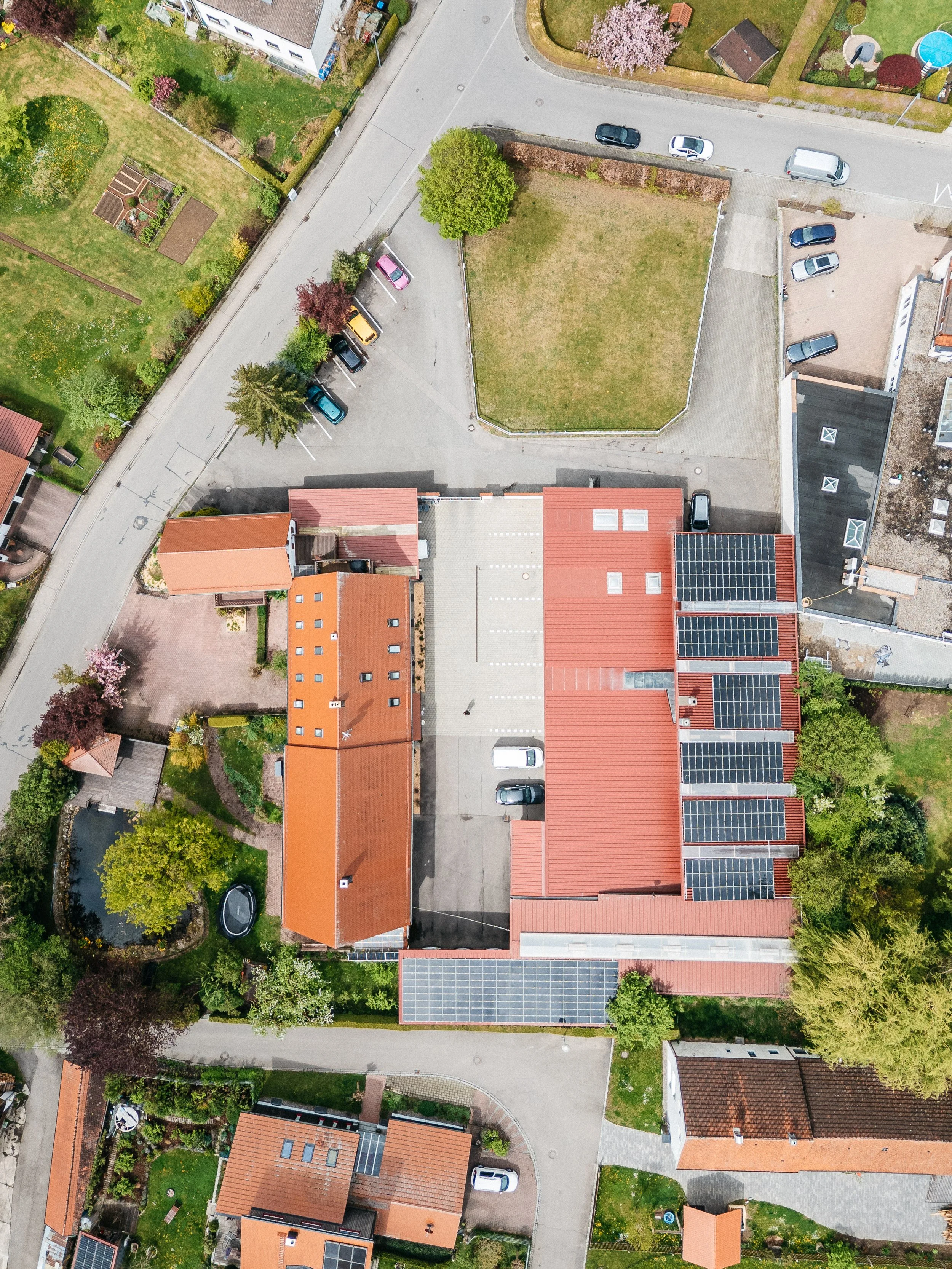Luftaufnahme eines Wohnviertels mit mehreren Gebäuden, Parkplätzen, Gärten und Bäumen. Die Gebäude haben rote Dächer, einige sind mit Solarpanelen versehen. Es gibt Fahrzeuge auf den Straßen und Parkplätzen.