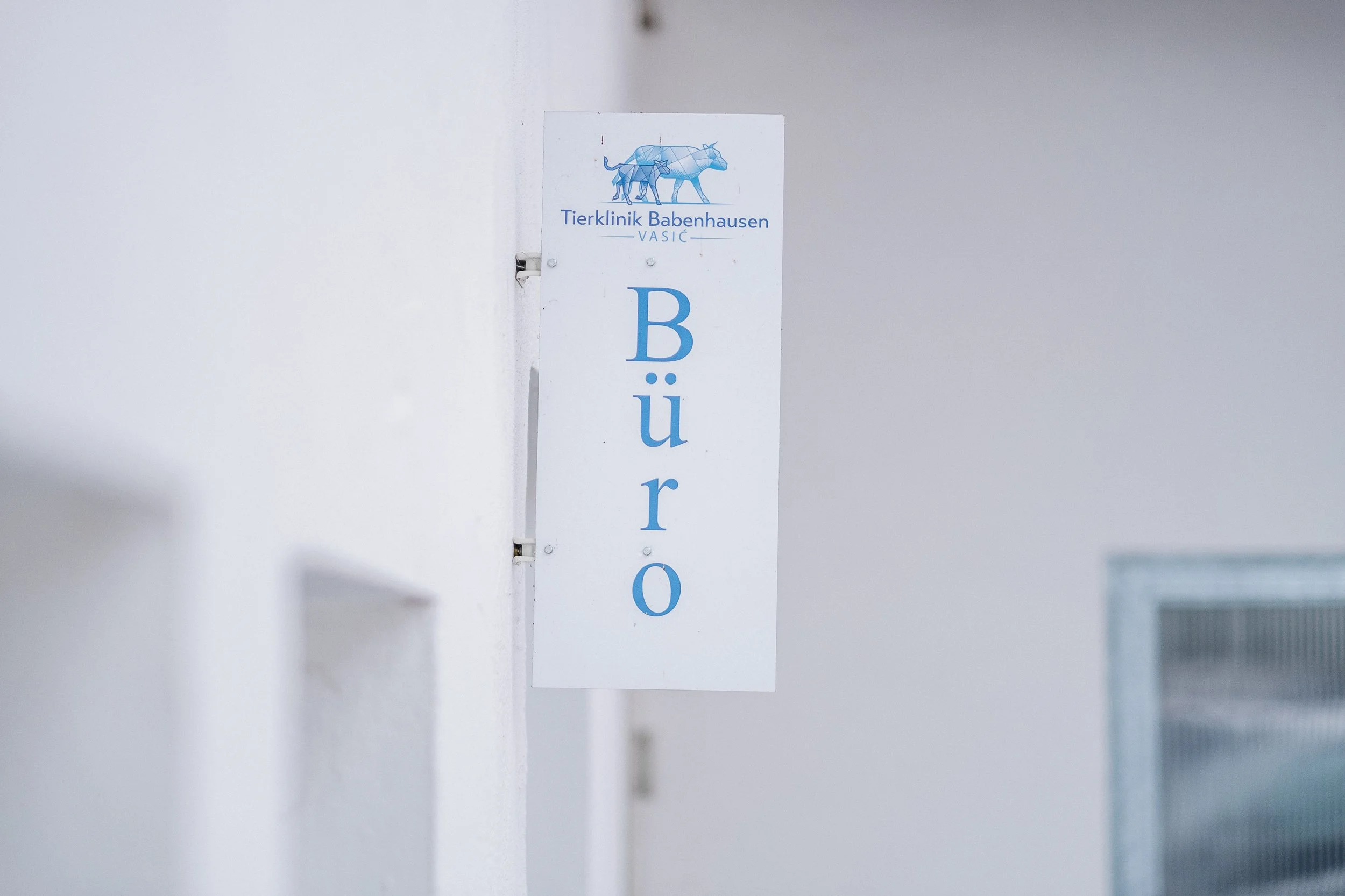 Ein weißes Schild mit blauer Schrift und einem Logo, das einen Hund und eine Katze zeigt, hängt an einer weißen Wand. Das Schild trägt die Aufschrift 'Tierklinik Babenhausen' und zeigt den Buchstaben 'Büro' vertical.