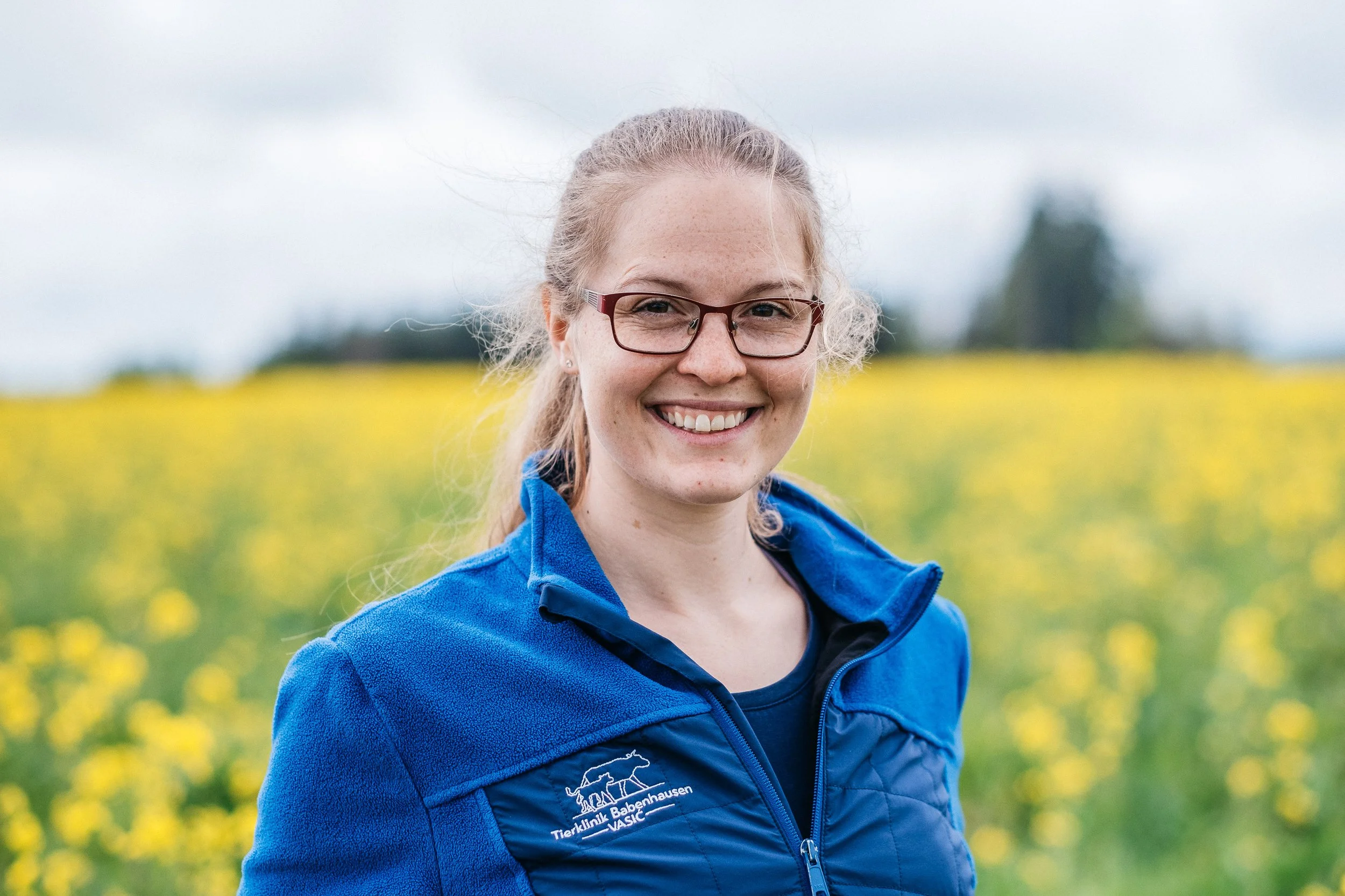 Lächelnde junge Frau mit Brille in blauer Jacke vor einem Blumenfeld