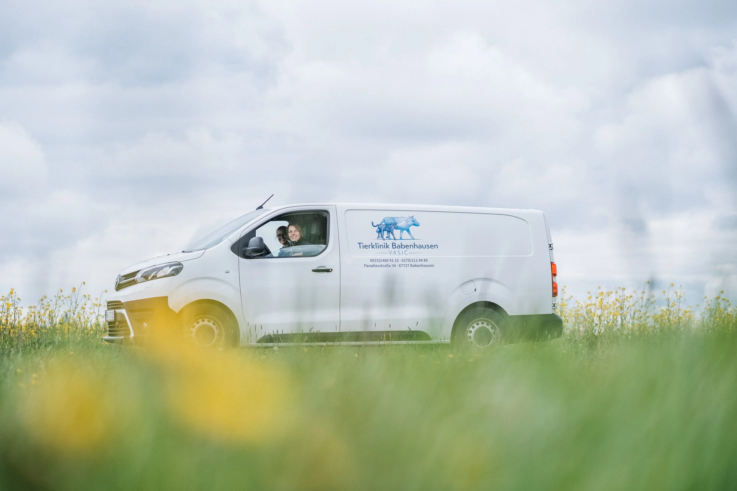Ein weißer Van mit Tierklinik-Logo und Text, parkend auf einer Wiese unter einem bewölkten Himmel, mit zwei lachenden Frauen im Fahrer- und Beifahrersitz.