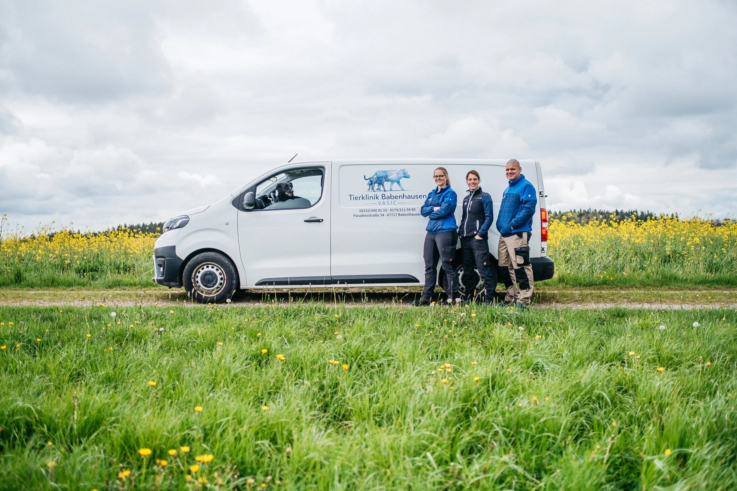 Drei Tierärzte mit einem weißen Lieferwagen vor einem Feld mit Mohnblumen, im Fahrzeug sitzt ein Hund, draußen stehen sie und lächeln in die Kamera.