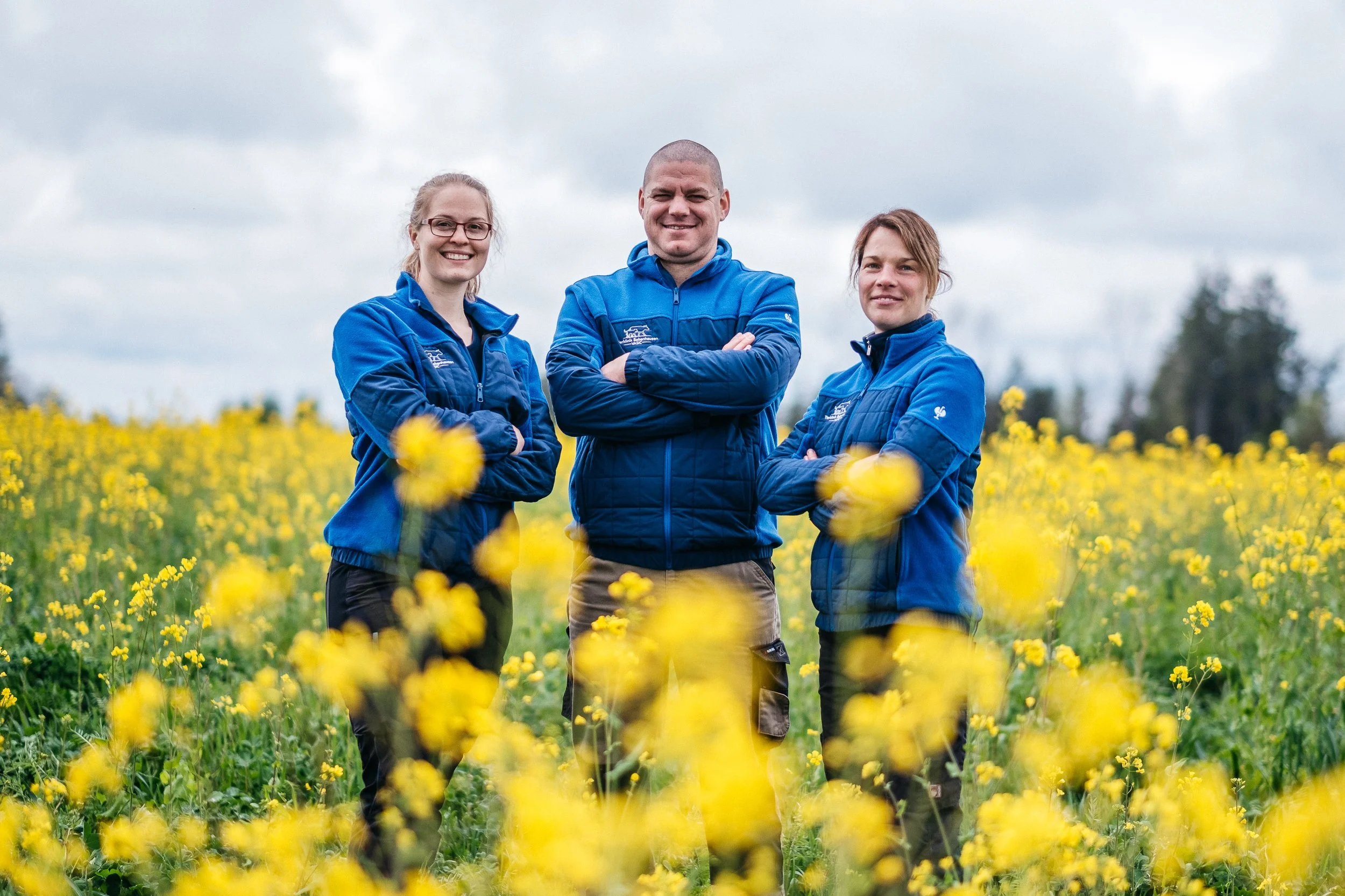 Drei Personen in blauen Jacken stehen im gelben Blumenfeld, lächeln und schauen in die Kamera, bei bewölktem Himmel.