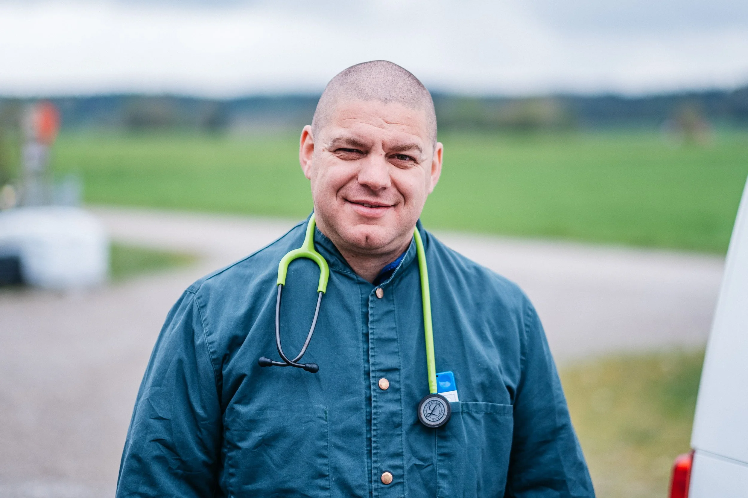 Ein männlicher Arzt trägt eine blaue Jacke und eine grüne Stethoskop um den Hals. Der Hintergrund ist eine offene, grüne Landschaft mit einem Weg.