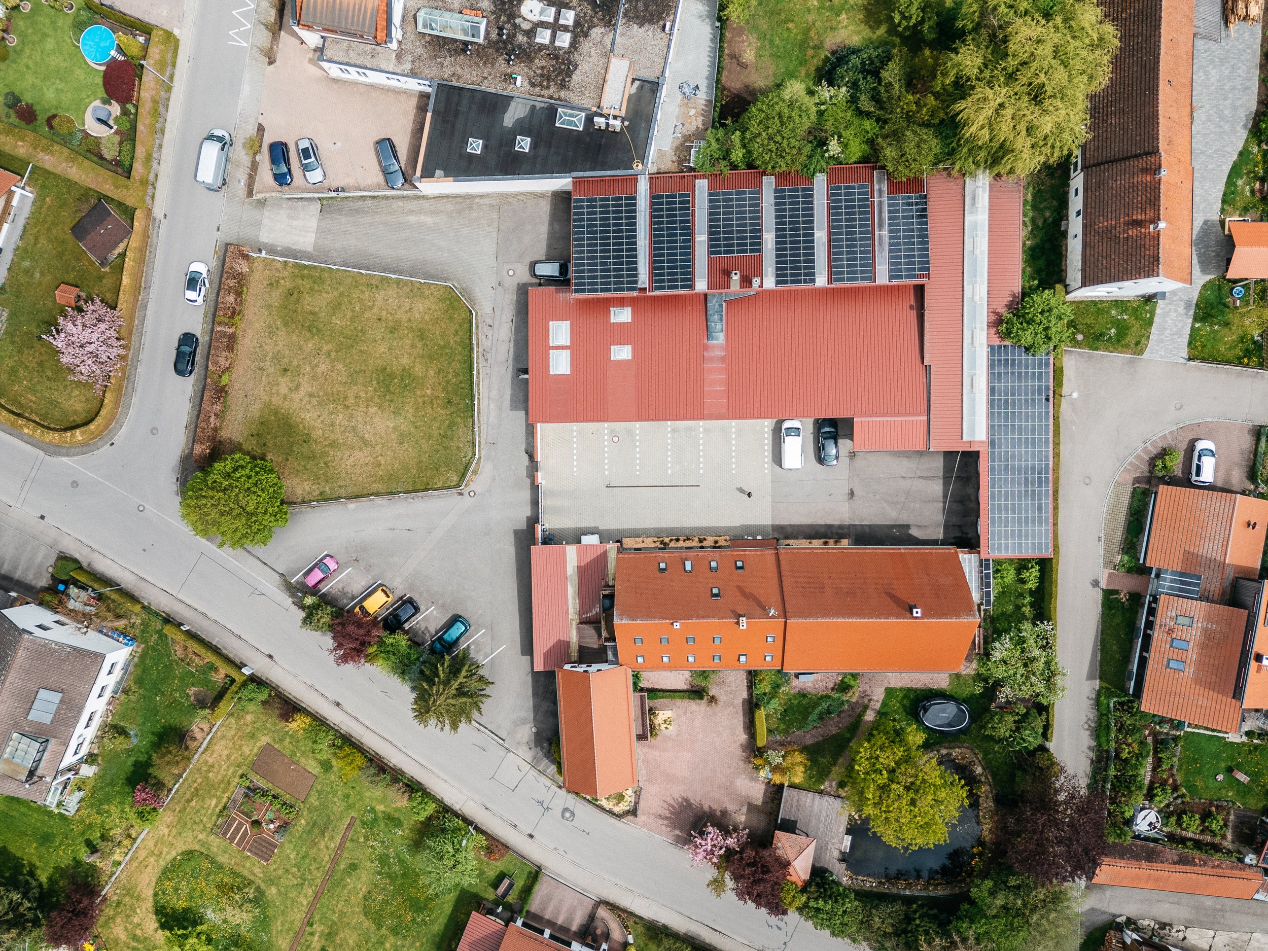 Luftaufnahme eines Gebäudes mit Solarpaneelen auf dem Dach, umgeben von Straßen, Parkplätzen, Gärten und Bäumen in einer wohnlichen Umgebung.
