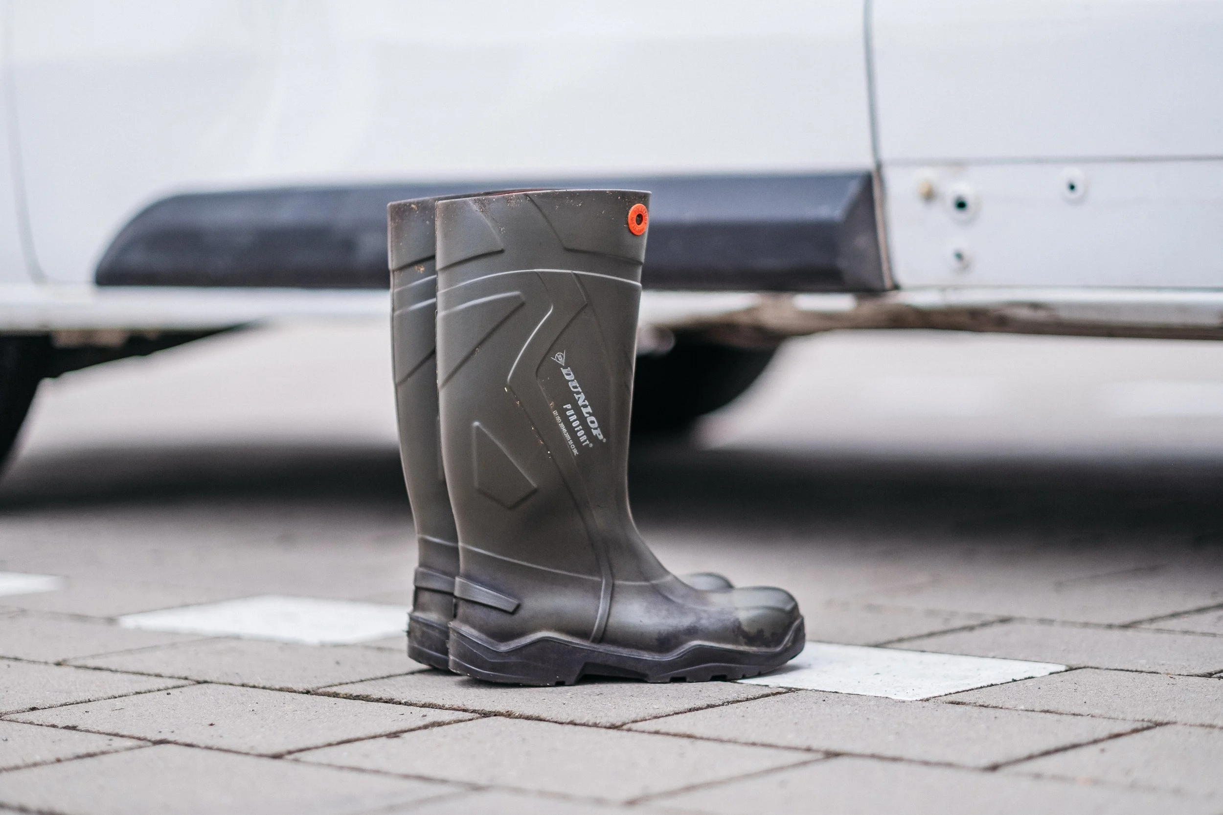 Ein schwarzer Gummistiefel steht auf einem Parkplatz auf weißen Markierungen, neben einem weißen Auto.