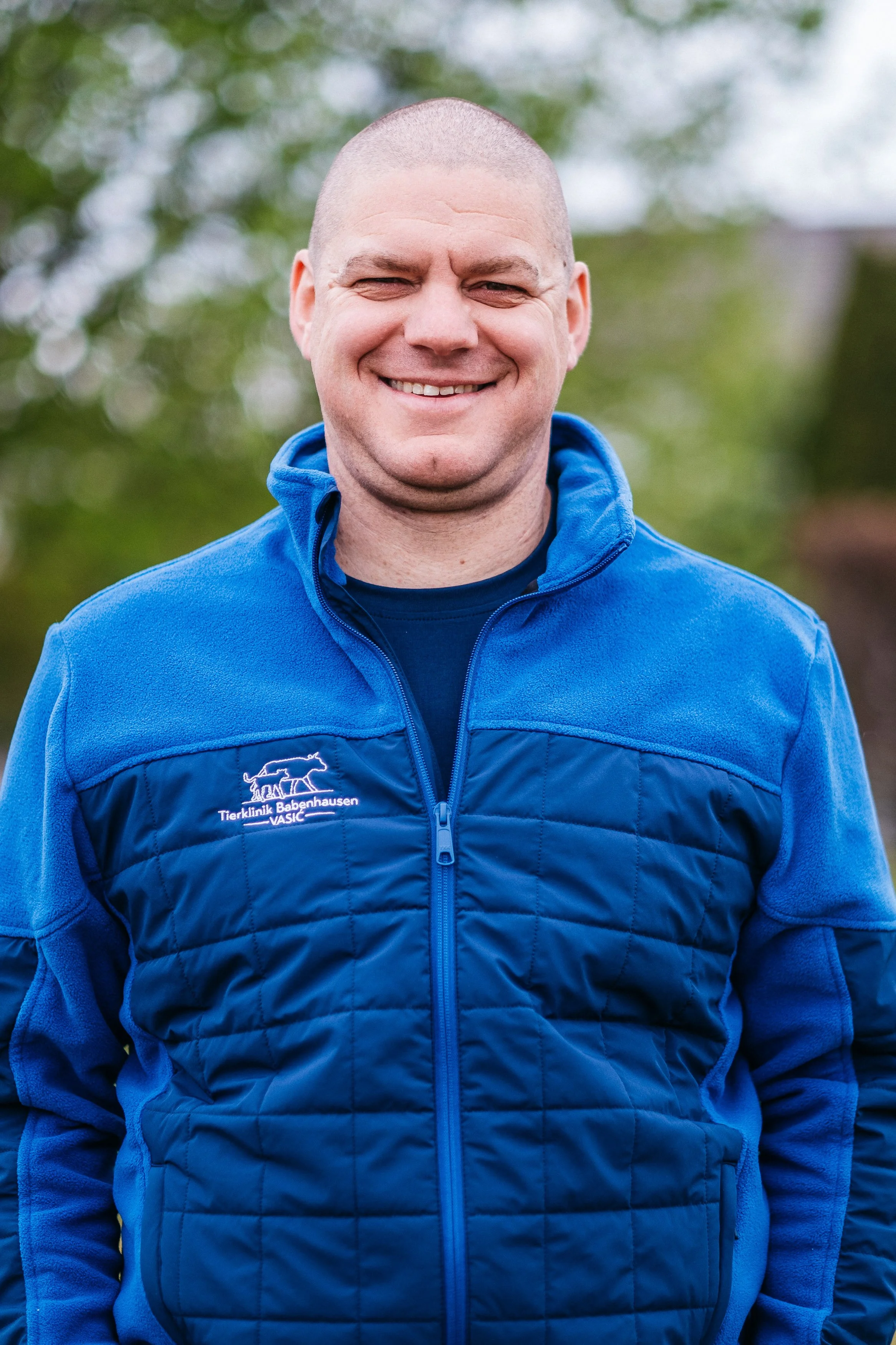 Ein lächelnder Mann trägt eine blaue Tierarztausrüstung mit dem Logo der Tierklinik Babenhausen auf der Brust, im Freien mit unscharfen grünen Bäumen im Hintergrund.