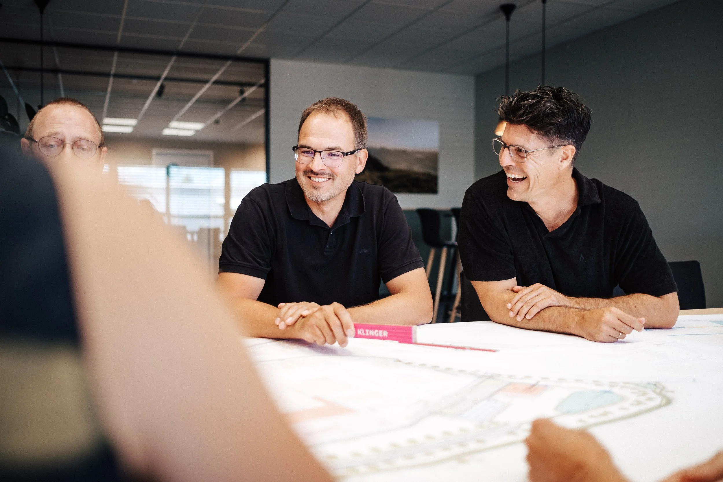 Drei Männer sitzen an einem Tisch und diskutieren, lachen und haben eine angenehme Unterhaltung in einem modernen Büro.